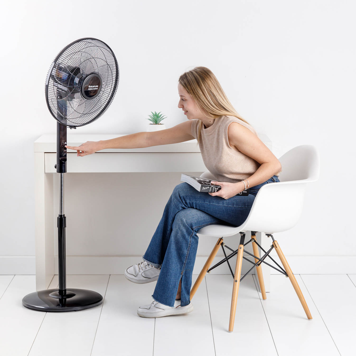 Una mujer ajusta un ventilador de pie negro cerca de un escritorio blanco. Está sentada en una silla blanca, sosteniendo un libro.