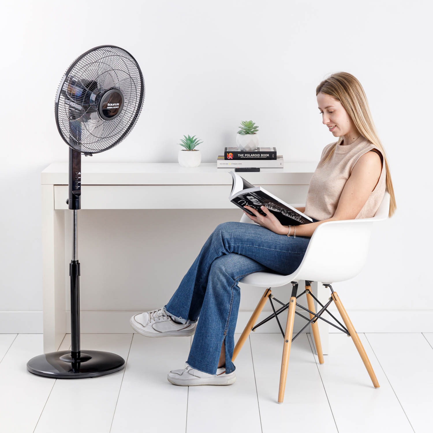 Mujer sentada, leyendo un libro, con un ventilador funcionando.