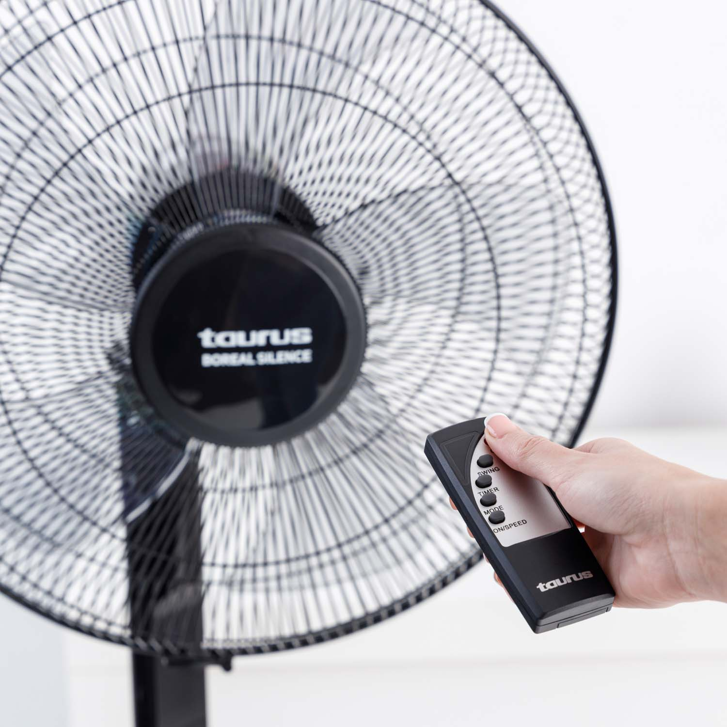 Un ventilador de pie negro con un mando a distancia en la mano. El ventilador está contra una pared blanca.