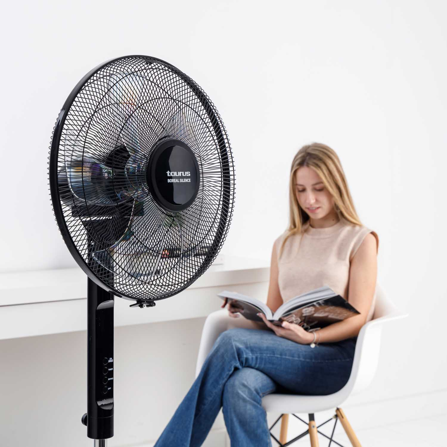 Una mujer leyendo un libro frente a un ventilador negro. El ventilador está sobre un soporte. Fondo blanco.