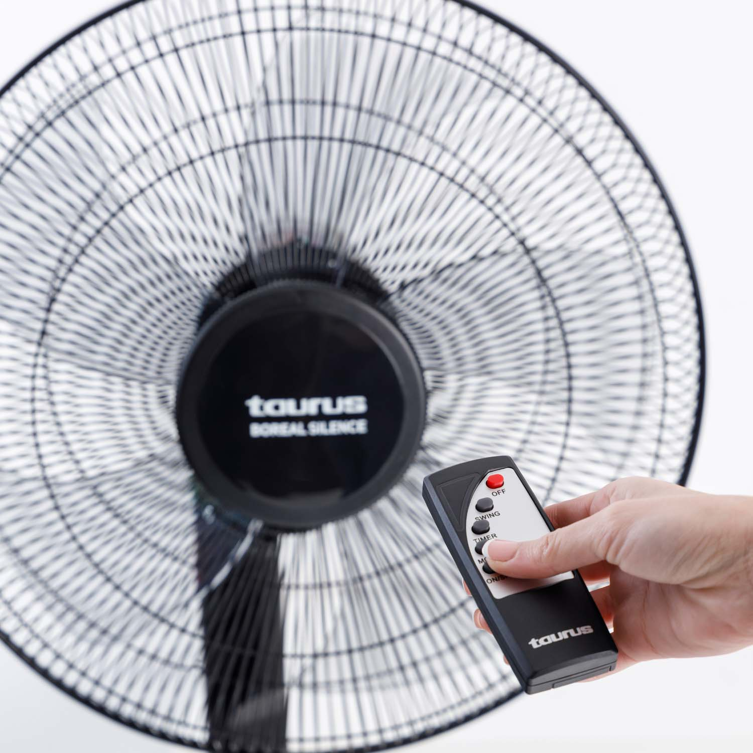 Una mano sostiene un mando a distancia para controlar un ventilador negro. El ventilador tiene un motor negro y una rejilla de malla metálica.