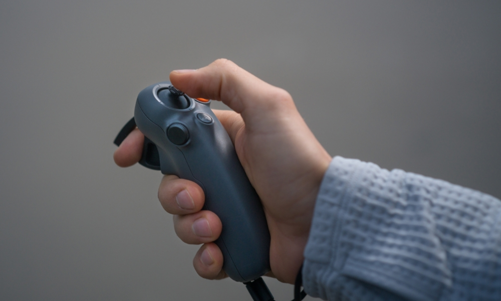 Dłoń trzymająca szary kontroler z przyciskami i joystickiem. Osoba ma na sobie szary sweter.