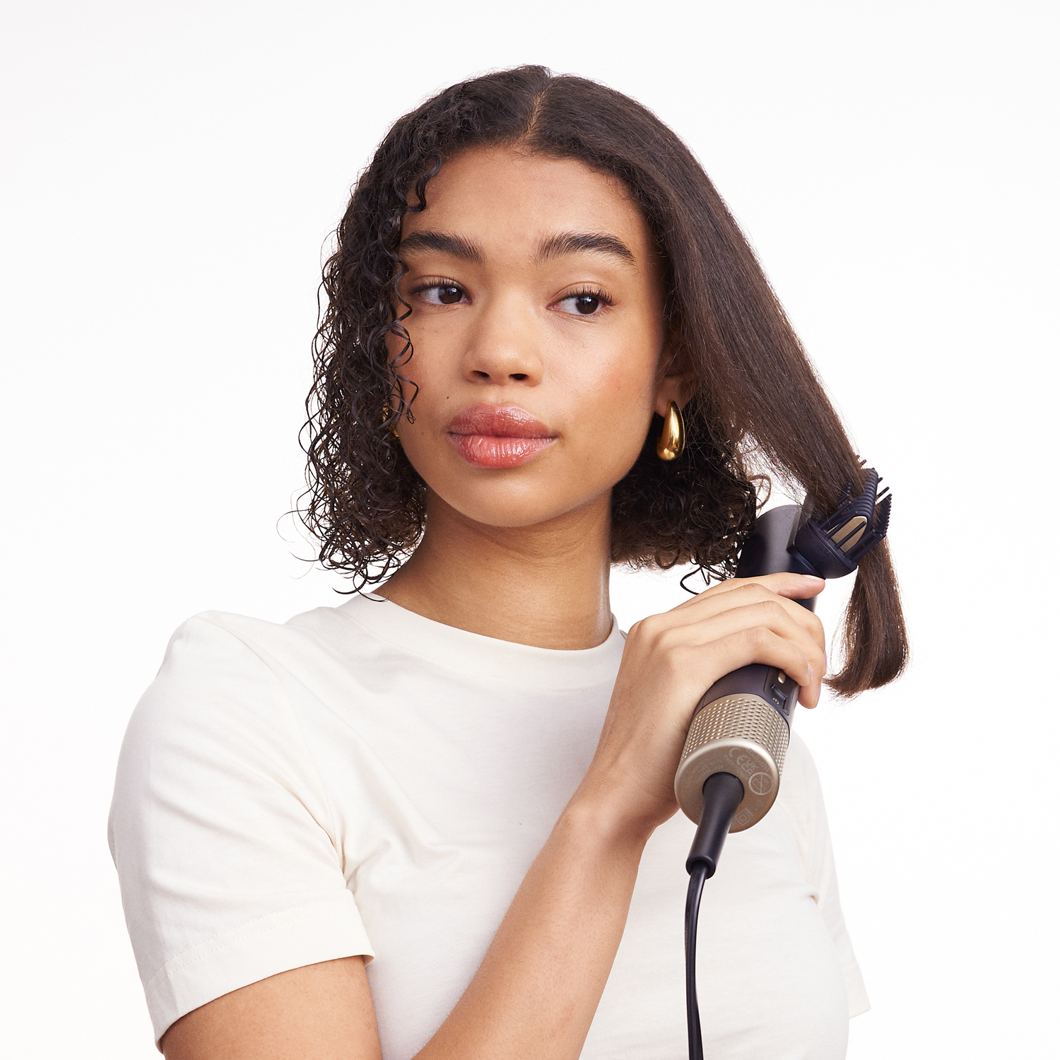 Woman using a hair styler. She has curly hair and wears a white shirt.