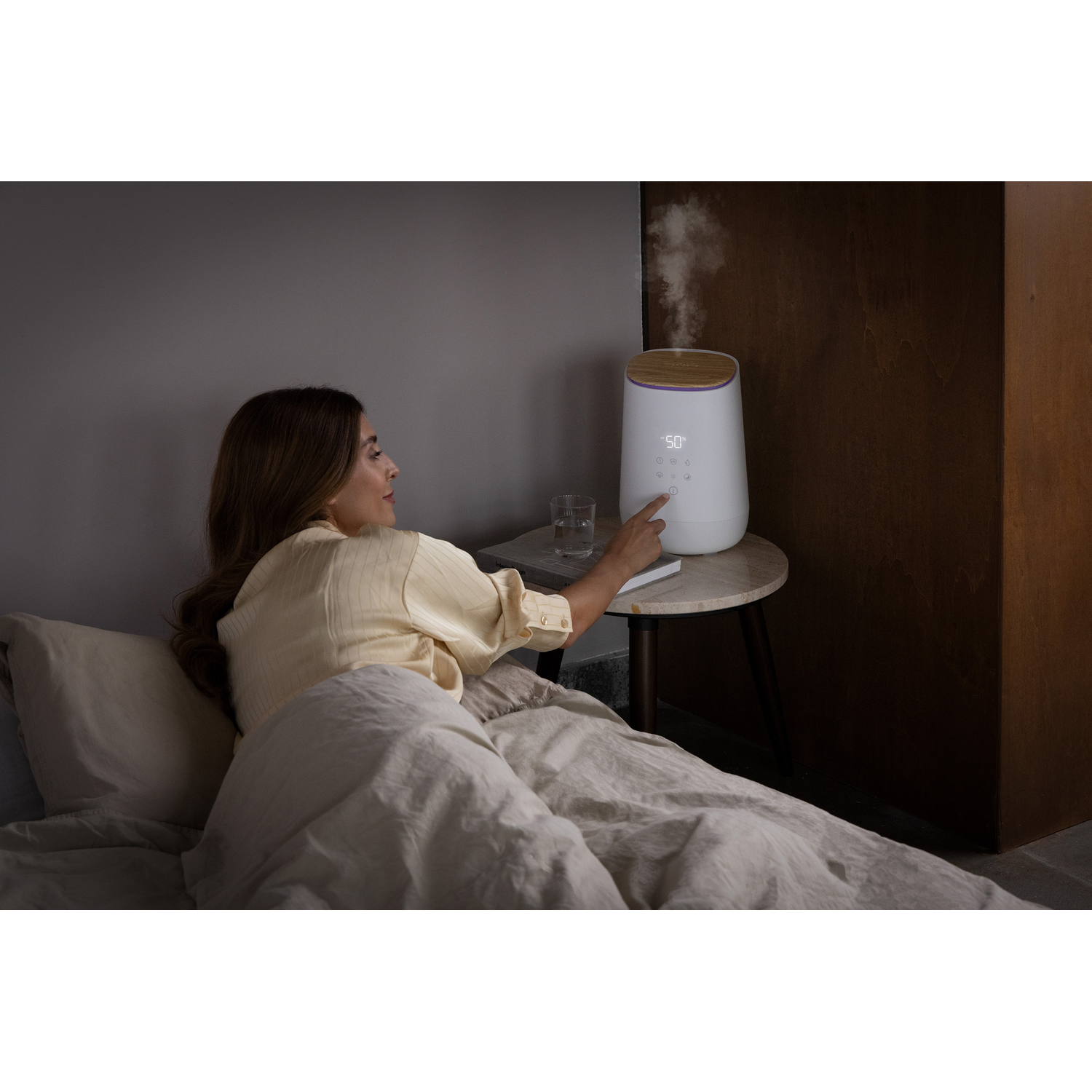 Vrouw bij een witte luchtbevochtiger op een tafel, met een glas water en een bed.