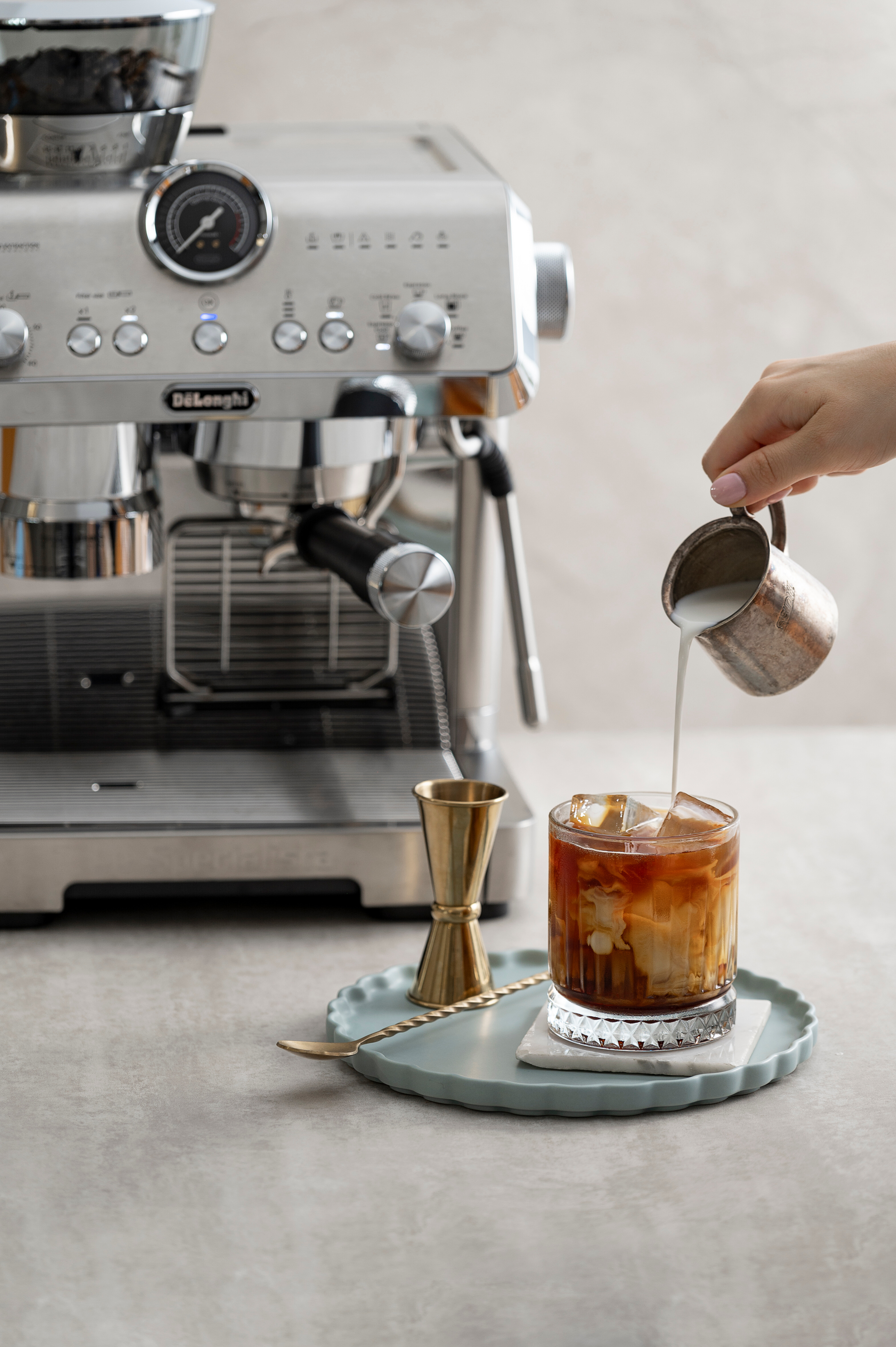 Eine Hand gießt Milch in ein Glas mit Eiskaffee. Eine De'Longhi Espressomaschine ist im Hintergrund.