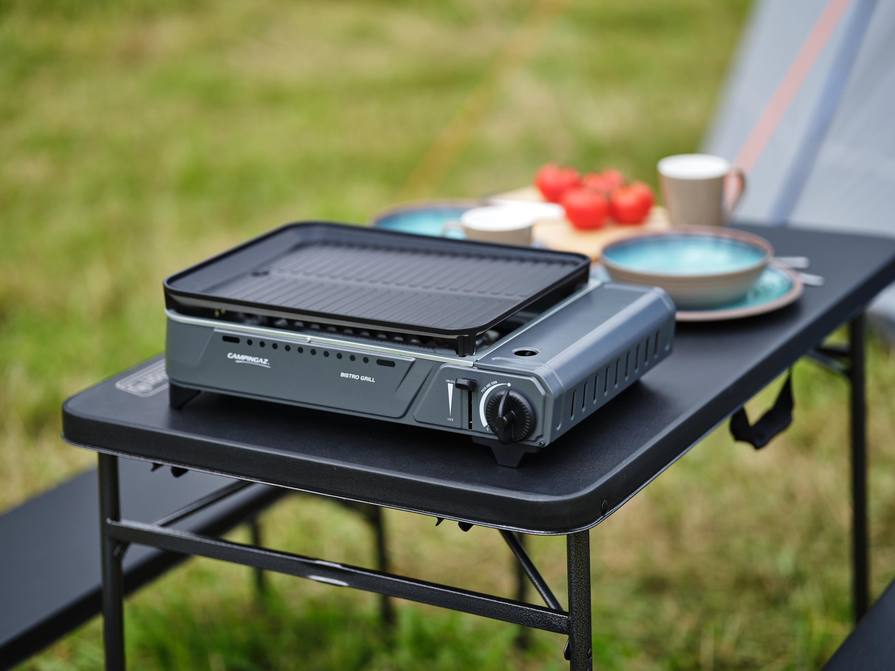Ein grauer Campingaz-Grill steht auf einem schwarzen Tisch mit Essen und Campingausrüstung auf einer Wiese.