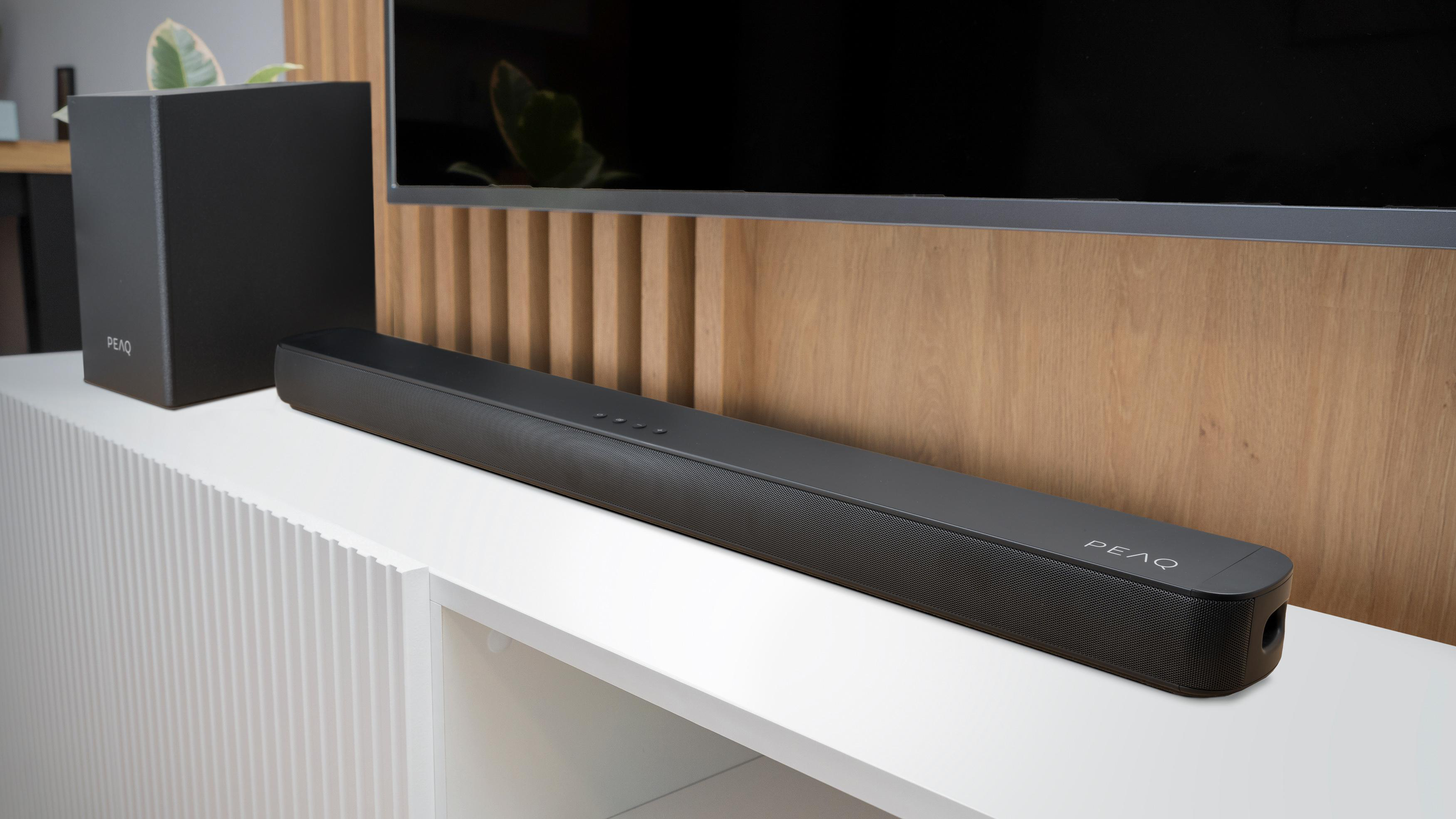 Eine schwarze Soundbar und ein Subwoofer auf einem weißen Schrank. Ein Fernseher ist im Hintergrund sichtbar.