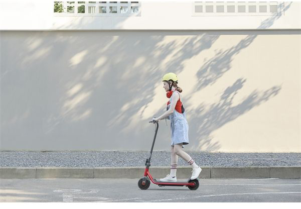 Egy lány piros robogóval közlekedik az utcán, sárga sisakkal és fejhallgatóval.