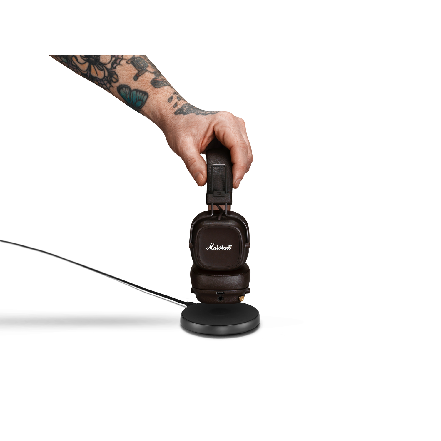 Een hand met tatoeages plaatst een Marshall-hoofdtelefoon op een oplaadstation. Witte achtergrond.