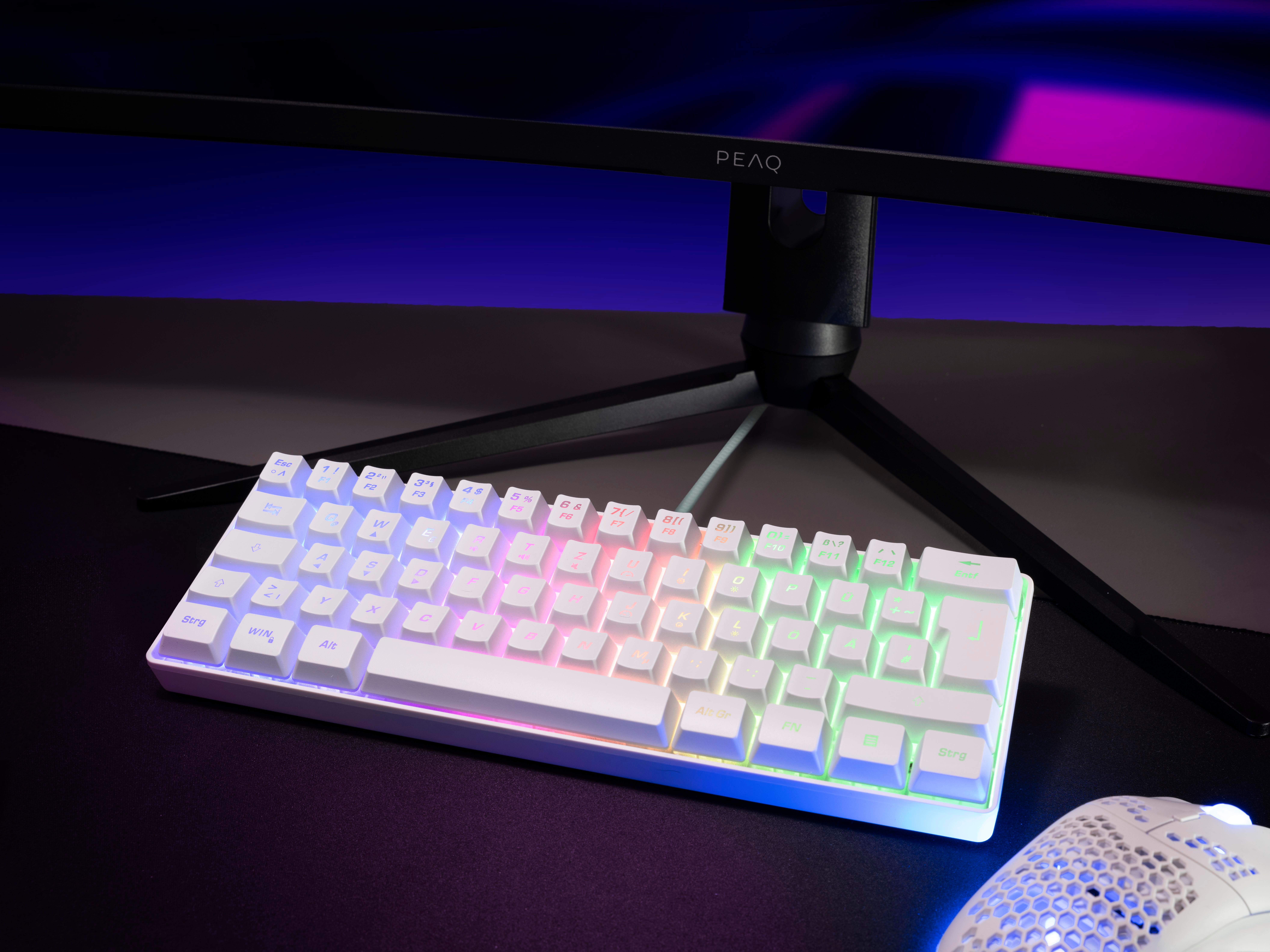 Weiße mechanische Tastatur mit farbenfroher Beleuchtung auf einer schwarzen Schreibtischunterlage, im Hintergrund ein Monitor.