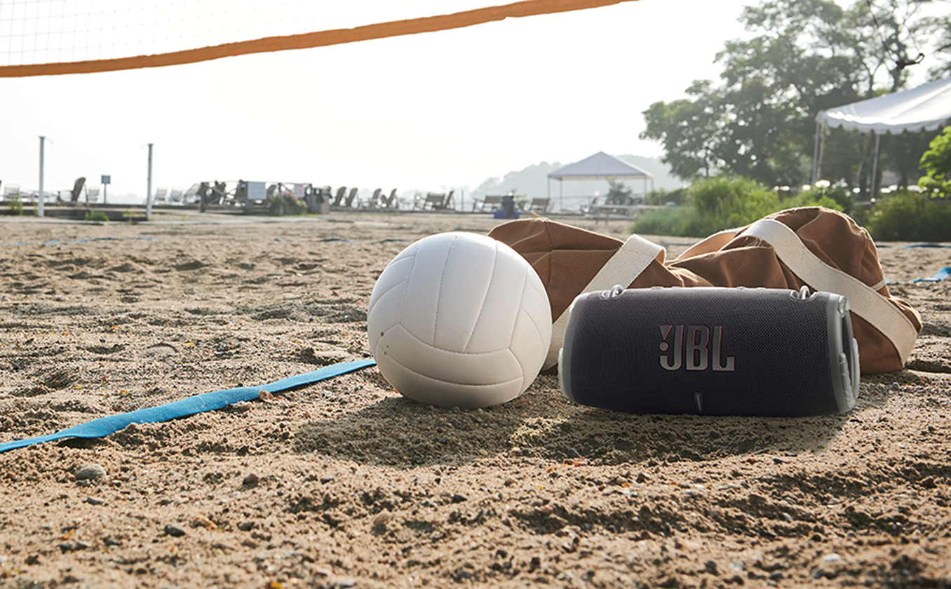 Strandjelenet: röplabda, táska, hangszóró, festői háttérrel, fákkal és vízzel.