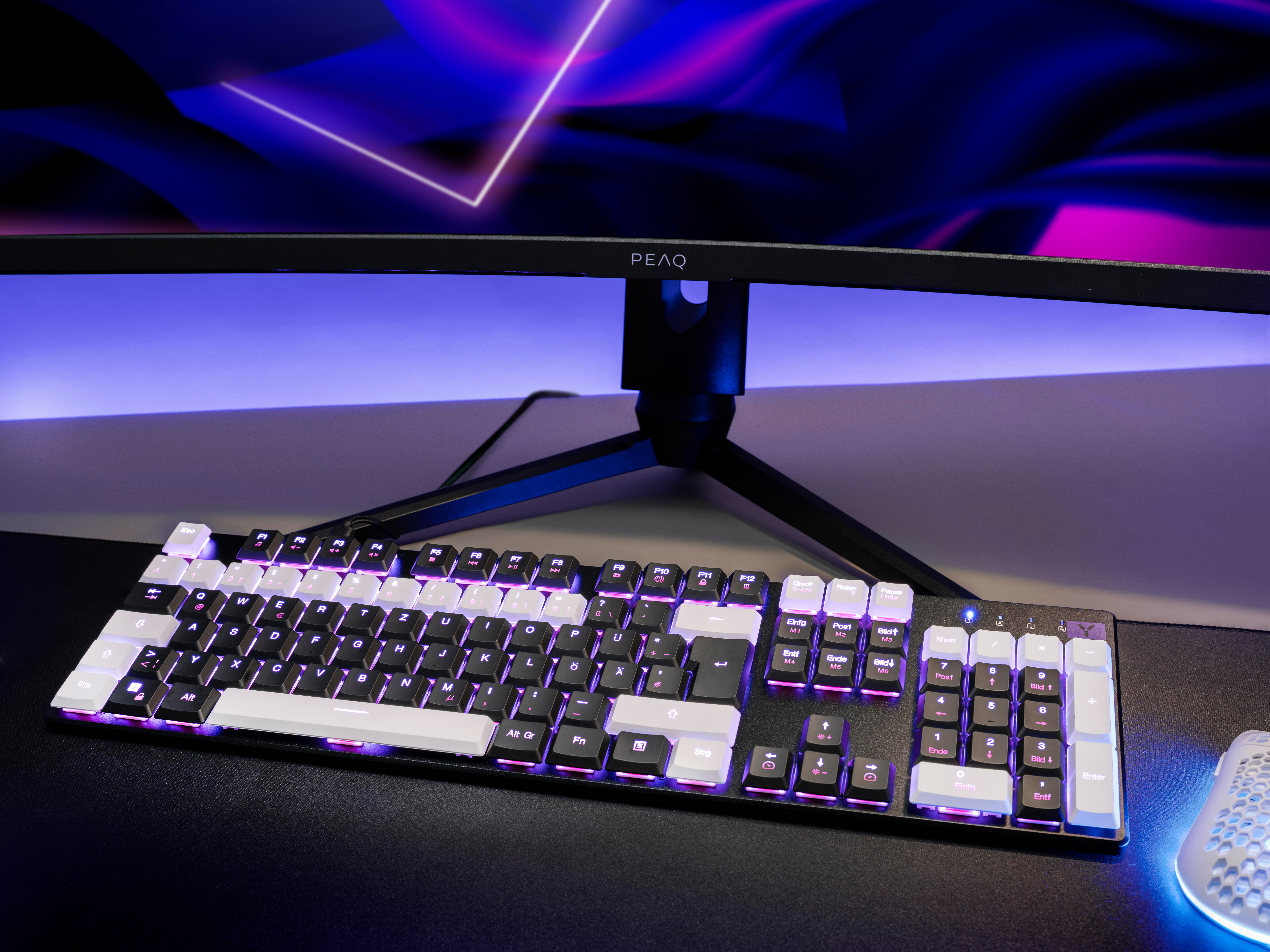 Schwarz-weiße Tastatur mit RGB-Beleuchtung und ein Monitor im Hintergrund.