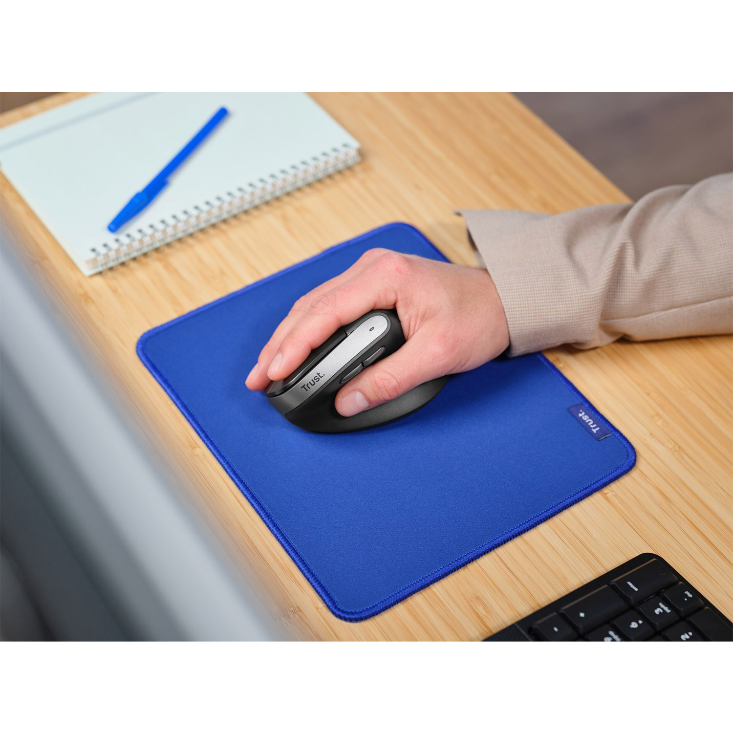 Una persona usa un mouse wireless su un tappetino blu su una scrivania in legno. Tastiera visibile, penna vicina.
