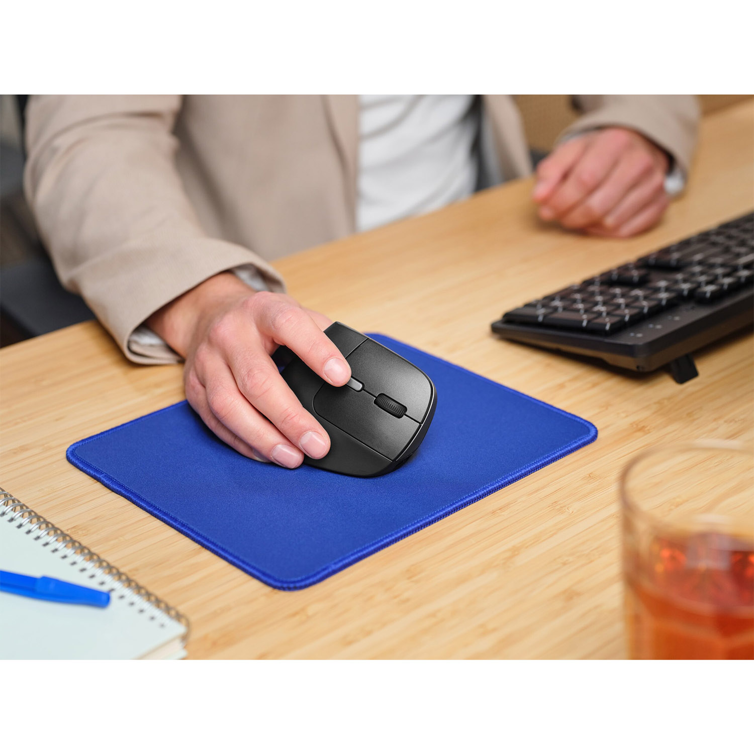 La mano di una persona che tiene un mouse nero su un tappetino blu. È visibile una tastiera.