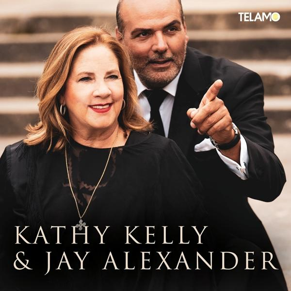 Kathy Kelly & Jay Alexander, ein Mann im Anzug zeigt, Frau lächelt, vor einer Backsteinmauer.