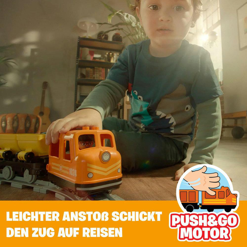 Ein Kind spielt mit einem orangefarbenen Spielzeugzug. Text sagt 'Push & Go Motor'.