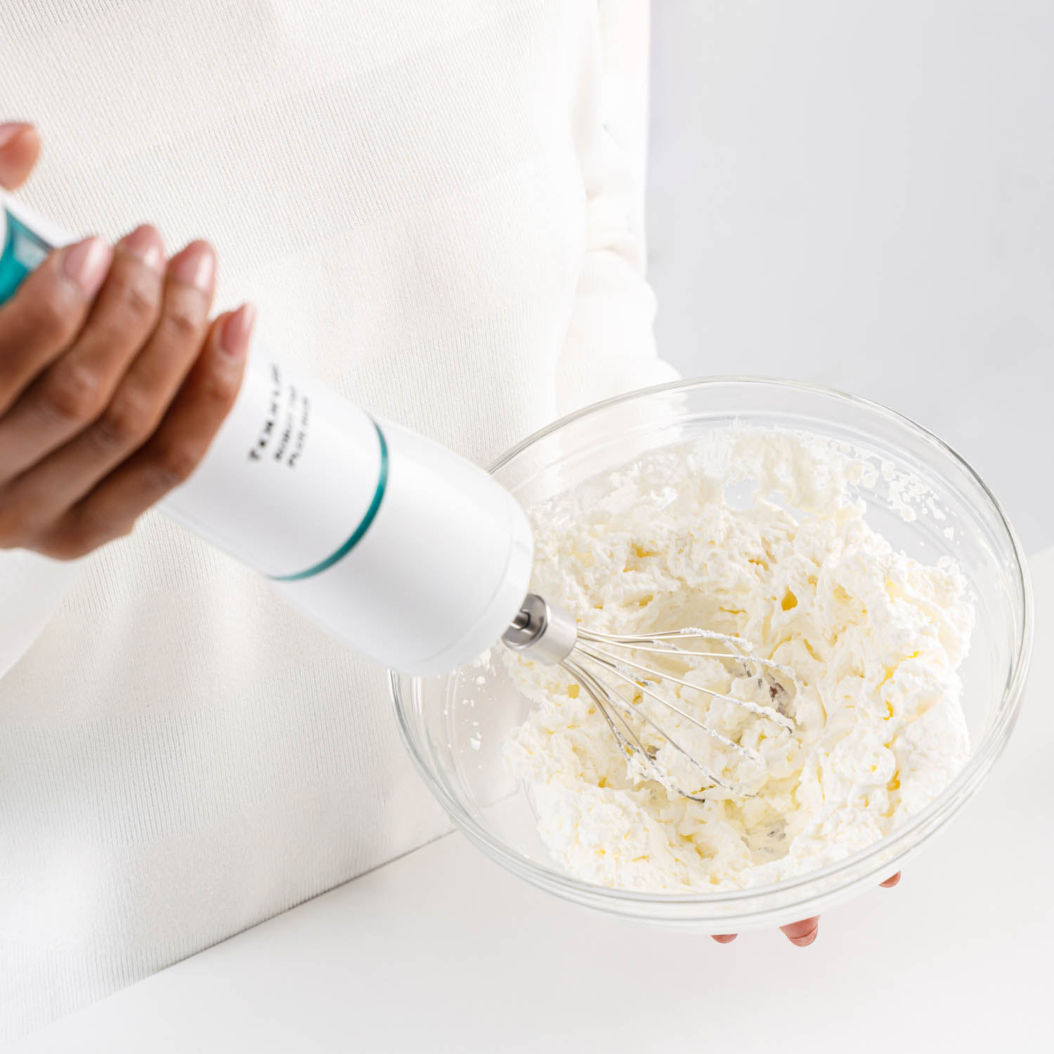 Una mujer usa una batidora de mano para mezclar crema en un bol de vidrio sobre una superficie blanca.