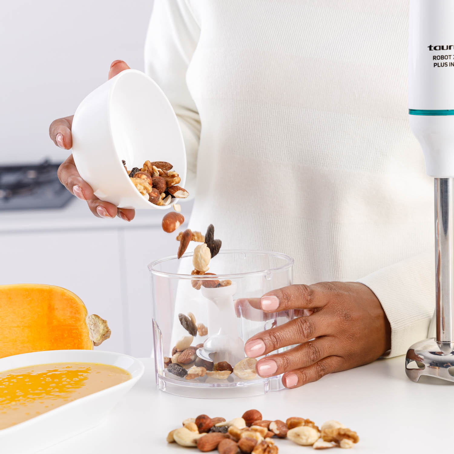 Una persona vierte nueces en un recipiente transparente, con una licuadora y un cuenco con nueces y calabaza al lado.