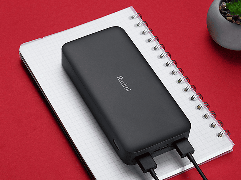 Fekete Redmi power bank egy fehér jegyzetfüzeten, piros felületre helyezve. Két fekete kábel csatlakozik.
