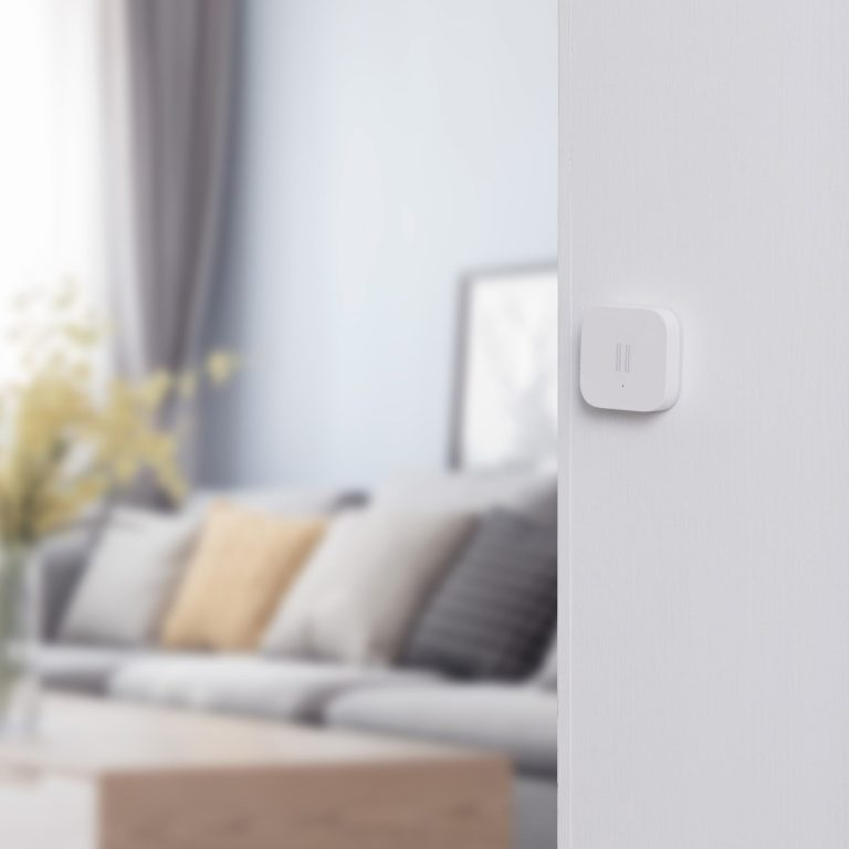 Un dispositivo per la casa intelligente bianco è su un muro bianco. Un'immagine sfocata di una stanza è sullo sfondo.