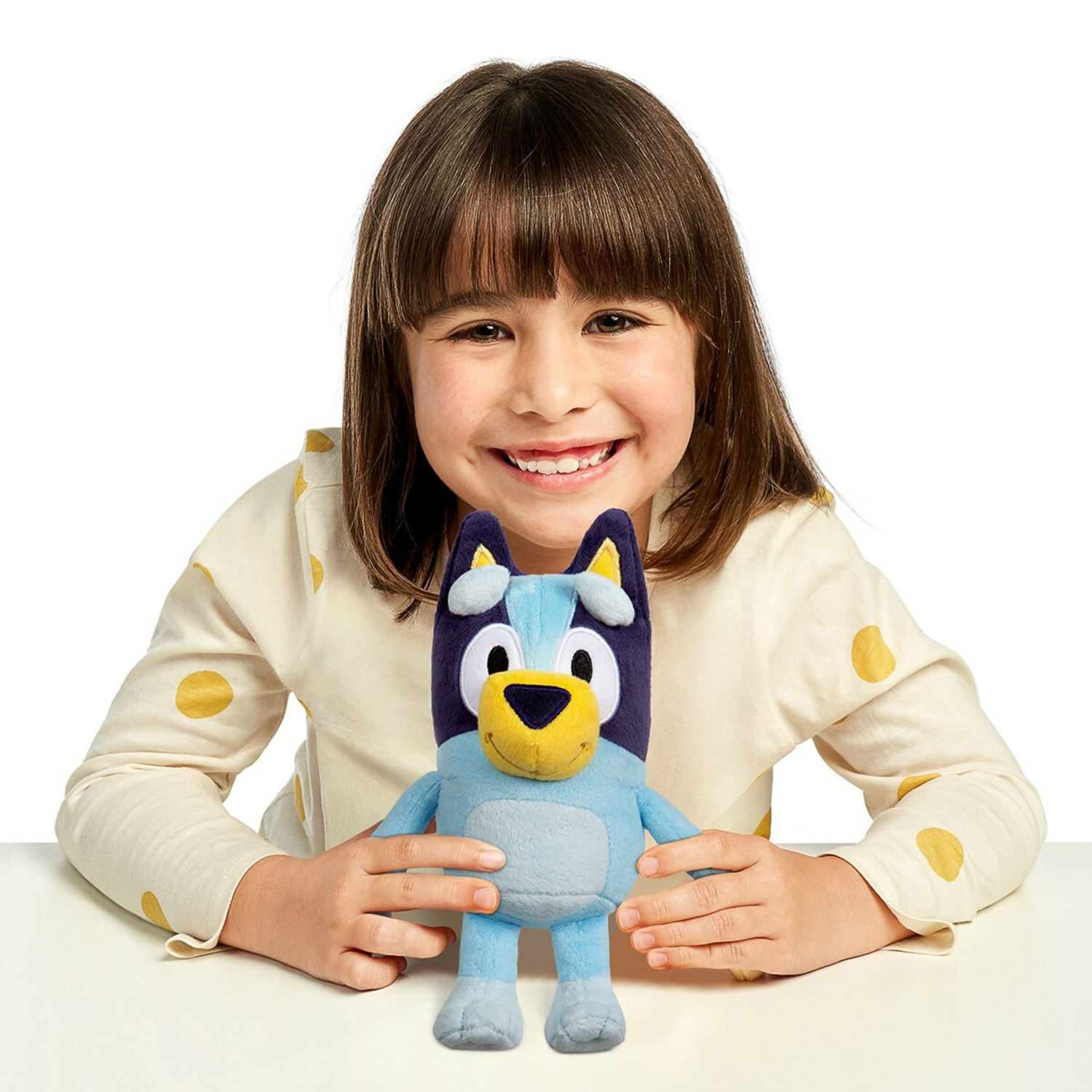 A smiling girl holding a Bluey plush toy. The girl wears a yellow shirt with white polka dots.