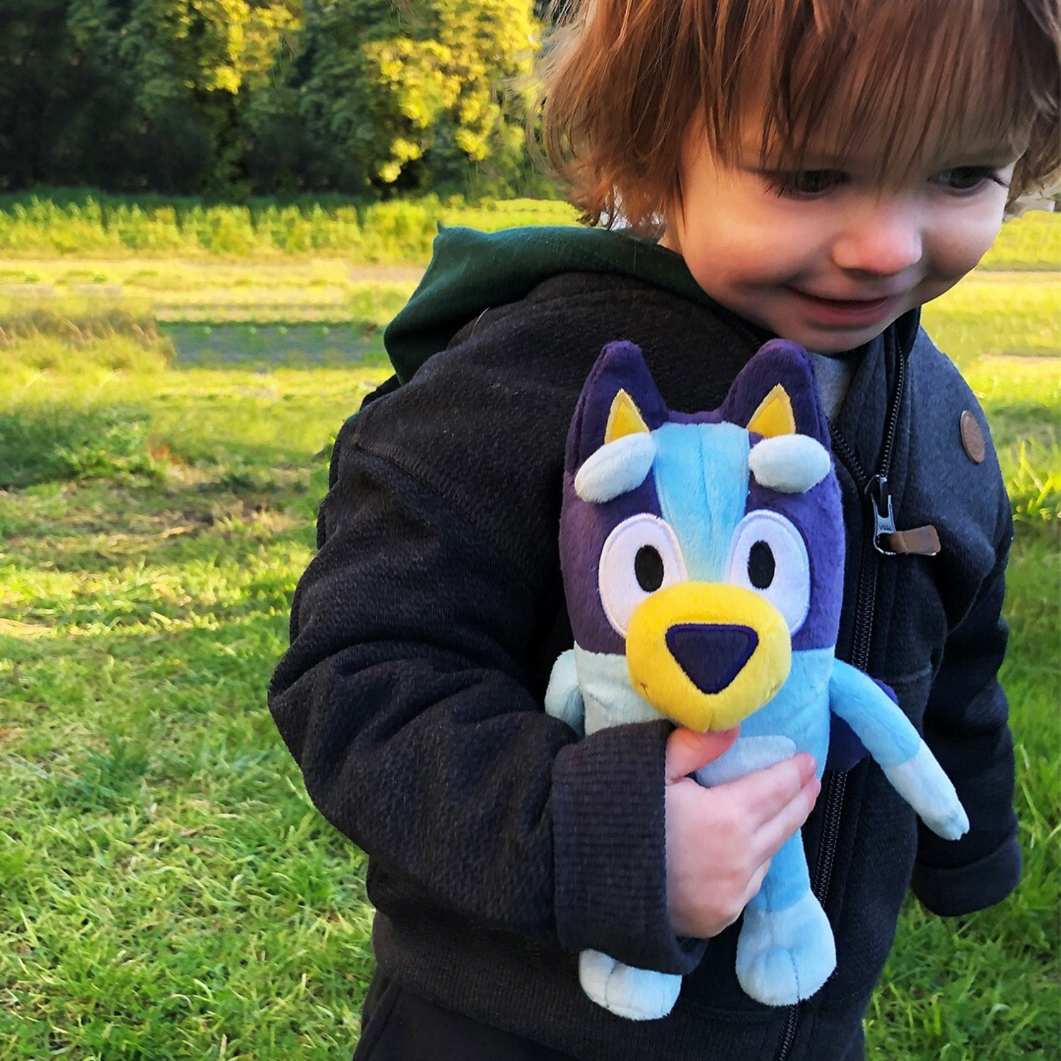A young child holding a blue plush toy. They are in a park with green grass and trees.
