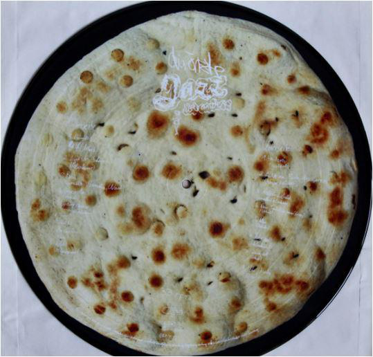 Pizzaförmige Schallplatte mit hellem Teig und dunklen Flecken. Das Etikett ist weiß, Text ist sichtbar.