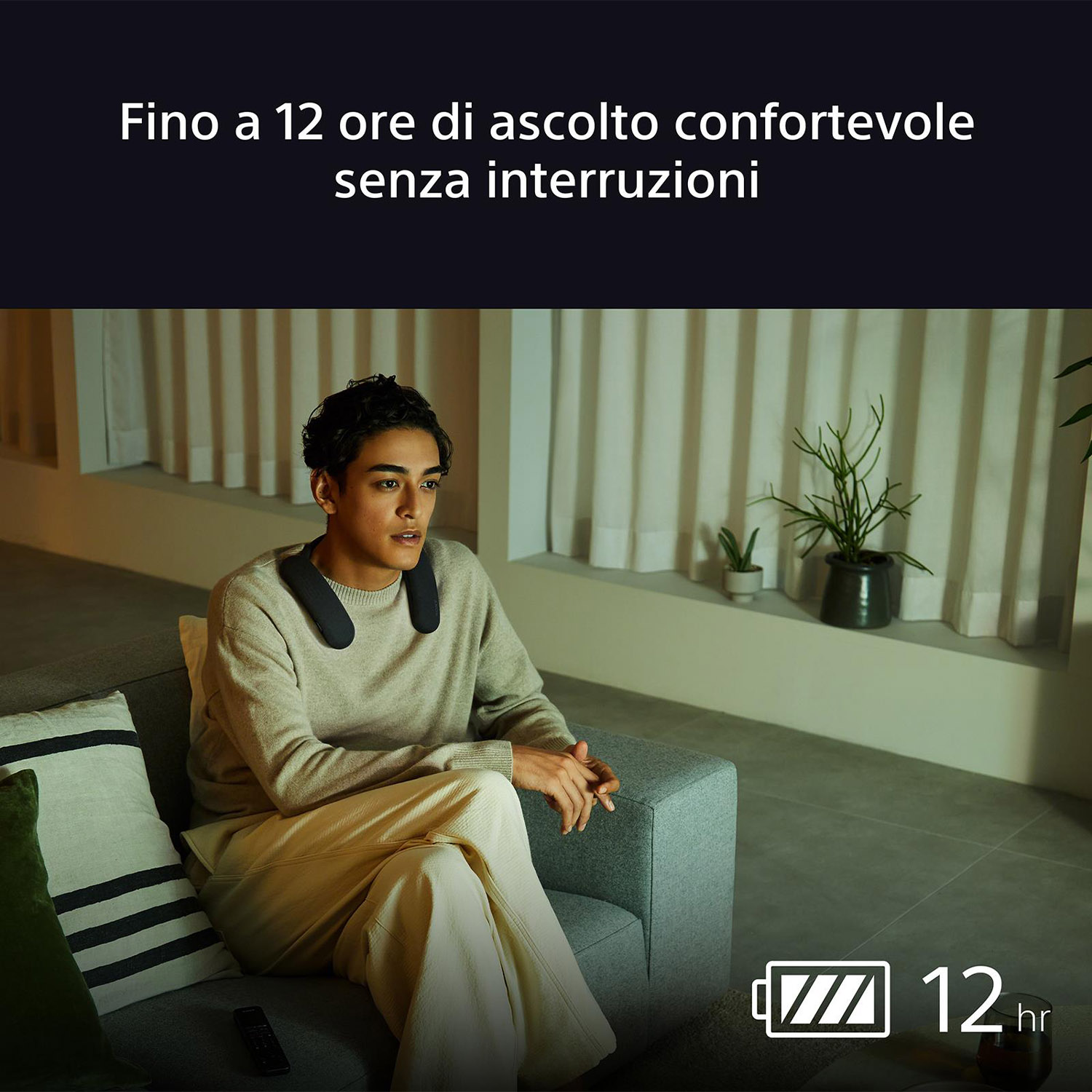 Una persona con un altoparlante da collo è seduta su un divano, che guarda qualcosa. Lo sfondo è una stanza buia.