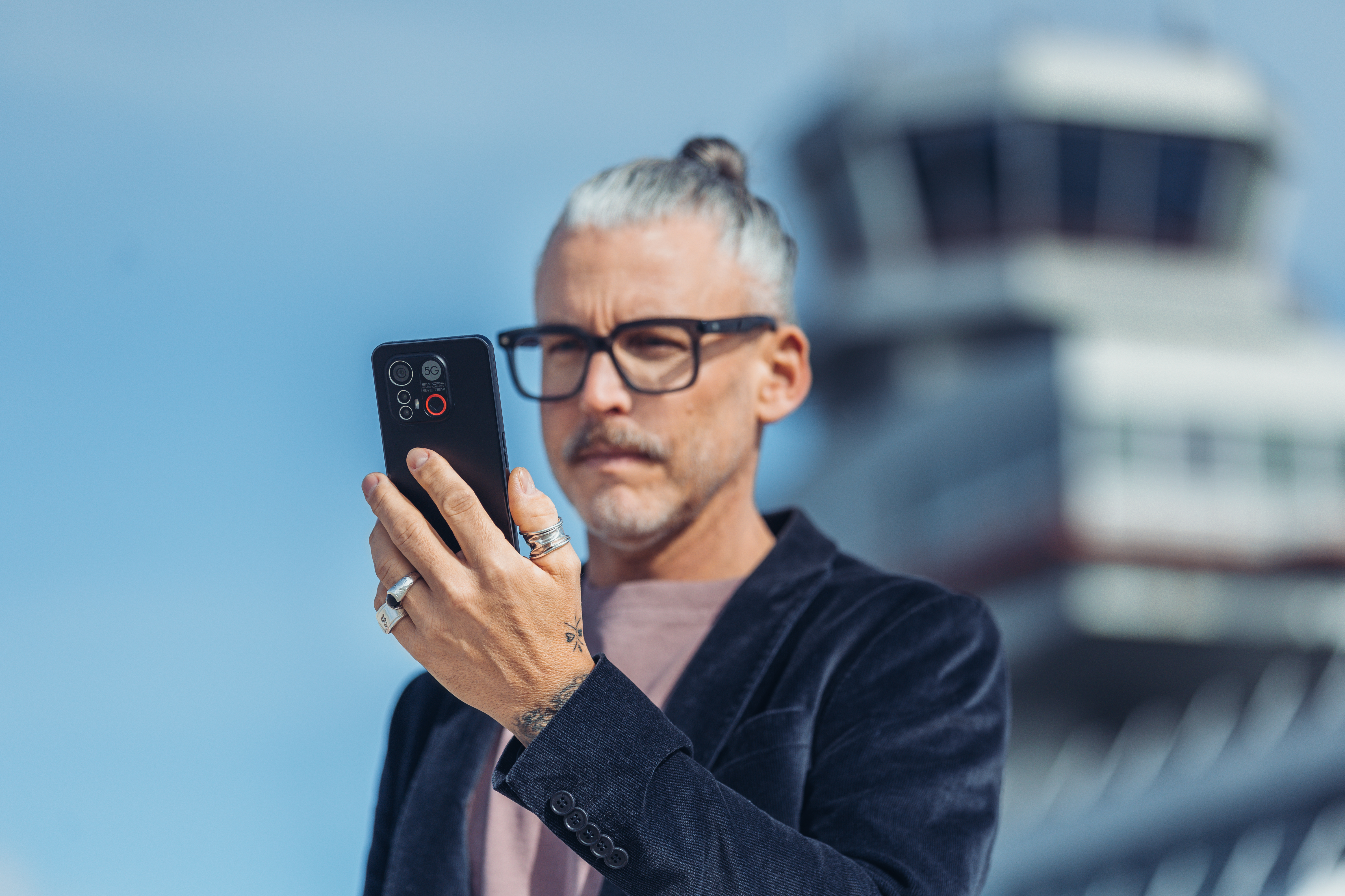 Ein Mann mit Dutt, Brille und Telefon, vor einem hohen Gebäude stehend.