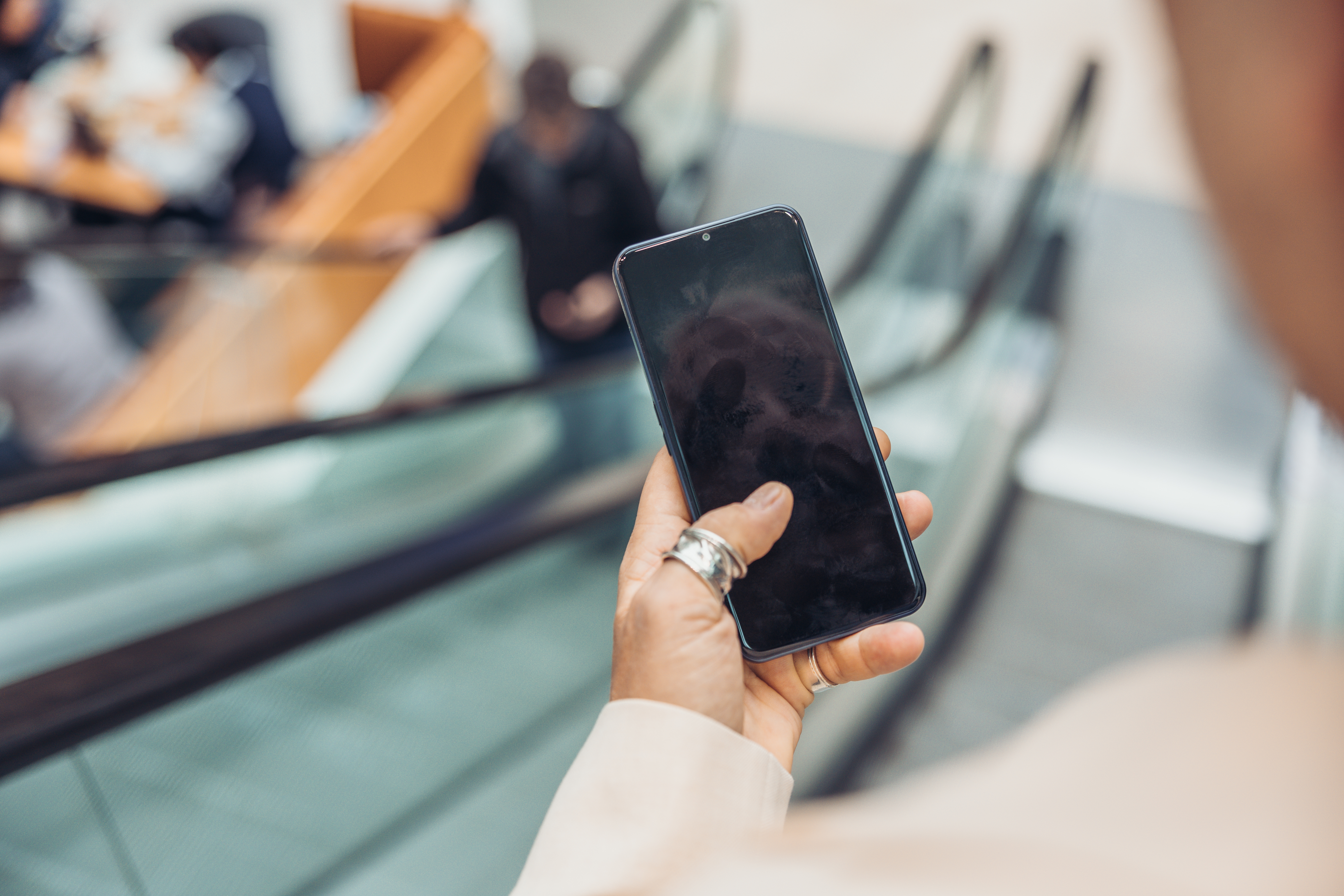 Person hält ein schwarzes Smartphone vor einer Rolltreppe. Der Bildschirm ist schwarz. Verschwommener Hintergrund.