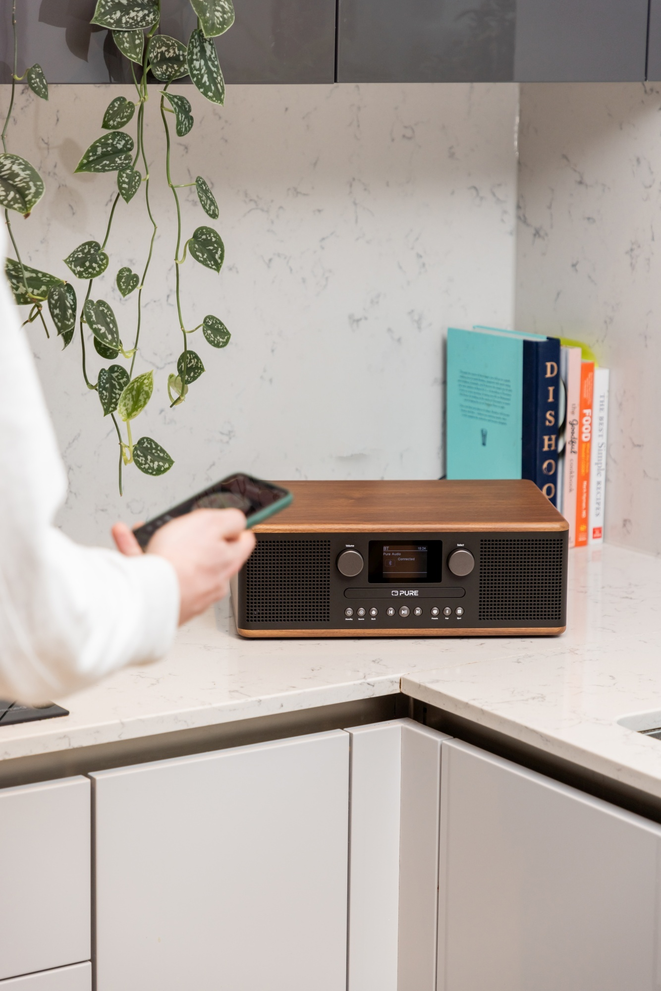 Une personne tient un téléphone près d'une radio en bois sur un comptoir. Livres et plantes en arrière-plan.
