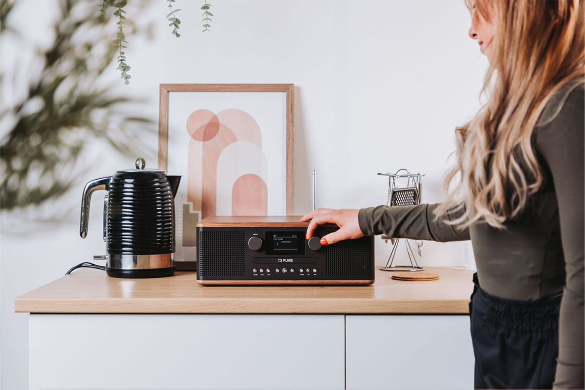 Femme réglant une radio sur un comptoir de cuisine. Bouilloire et œuvre d'art visibles. Plan de travail en bois.