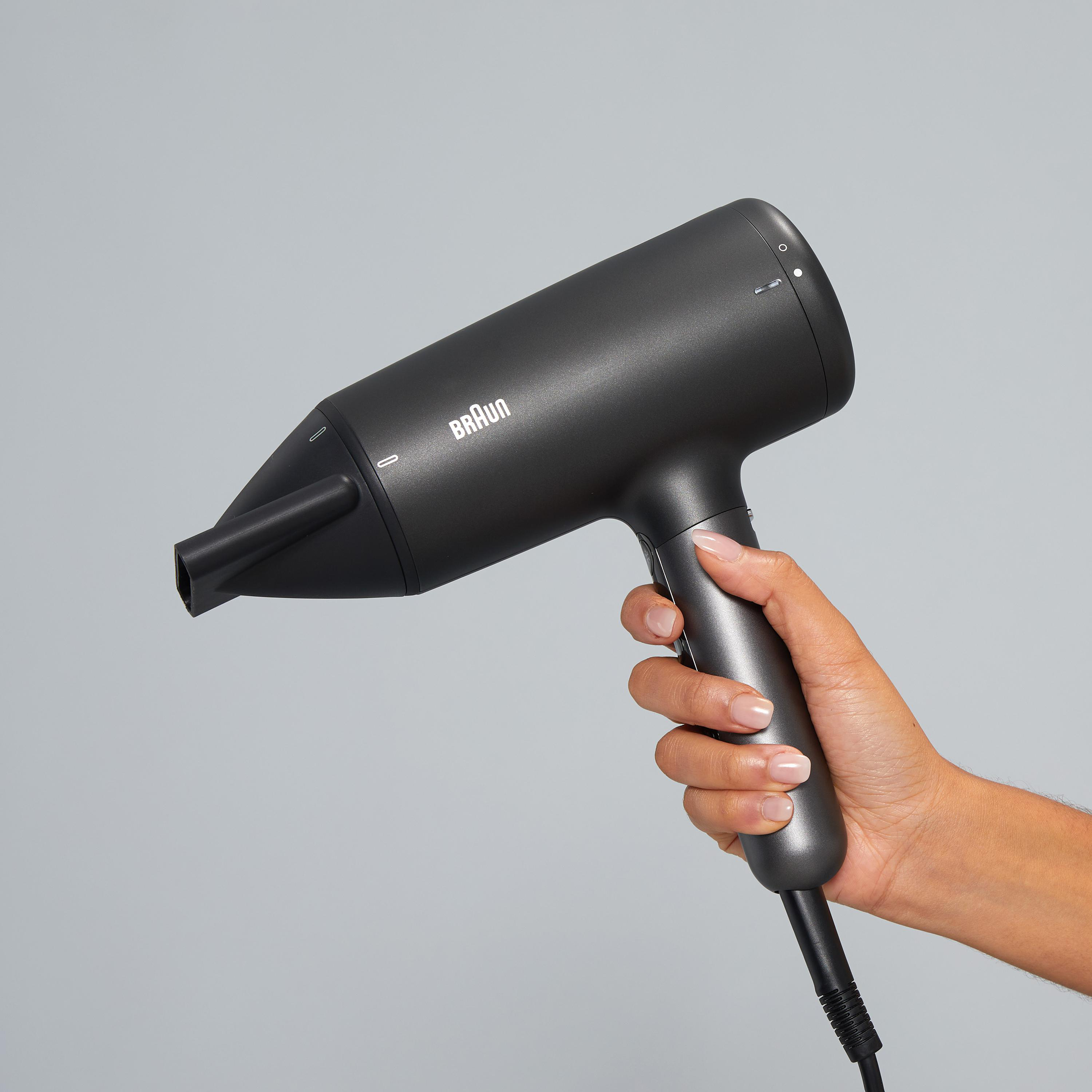 Ein schwarzer Braun Haartrockner wird in einer Hand vor helgrauem Hintergrund gehalten.
