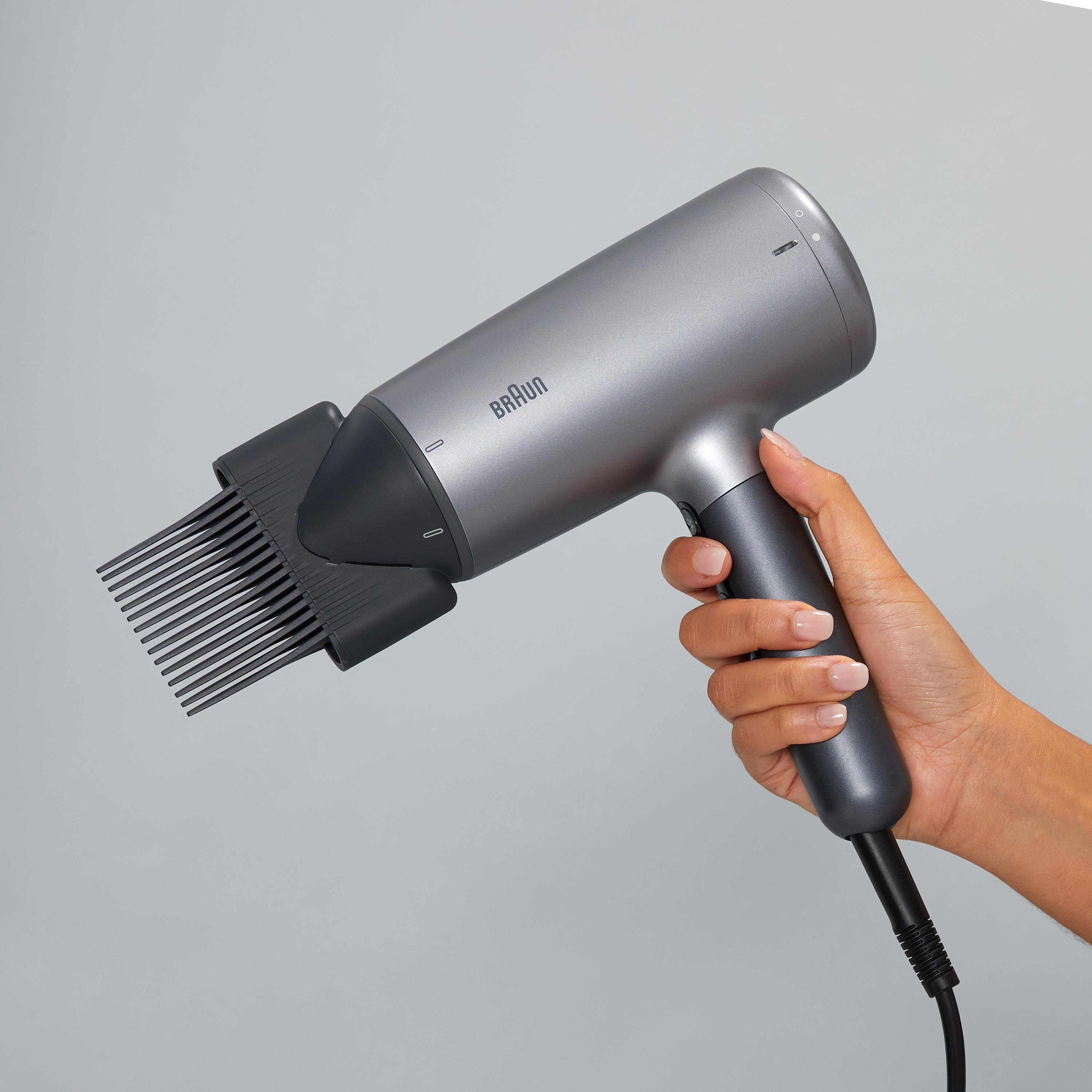 Eine Hand hält einen grauen Braun Haartrockner mit Kammaufsatz, auf grauem Hintergrund.