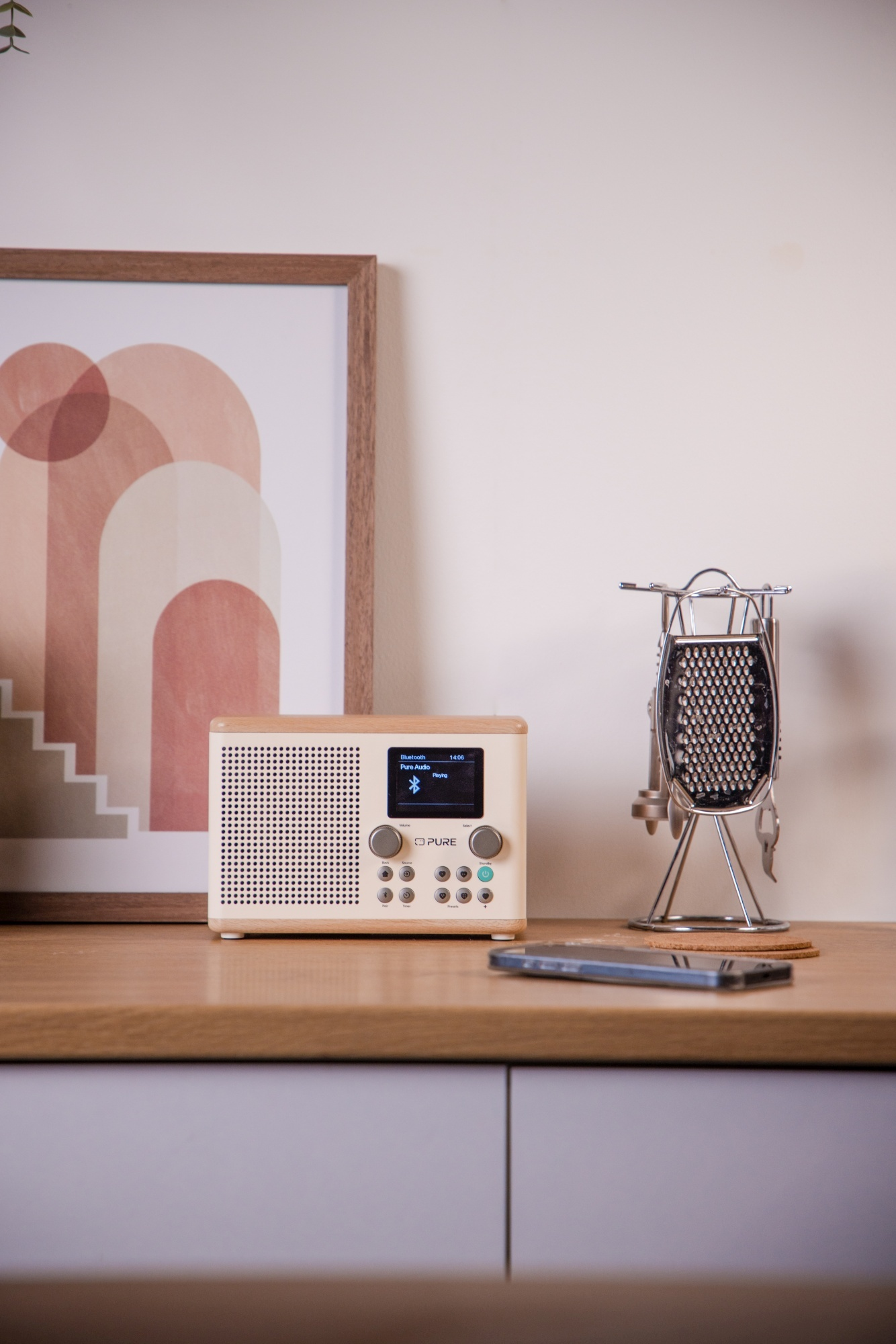 Ein Radio und ein Handy auf einem Holztisch. Ein Bild und eine Reibe befinden sich hinter dem Radio.