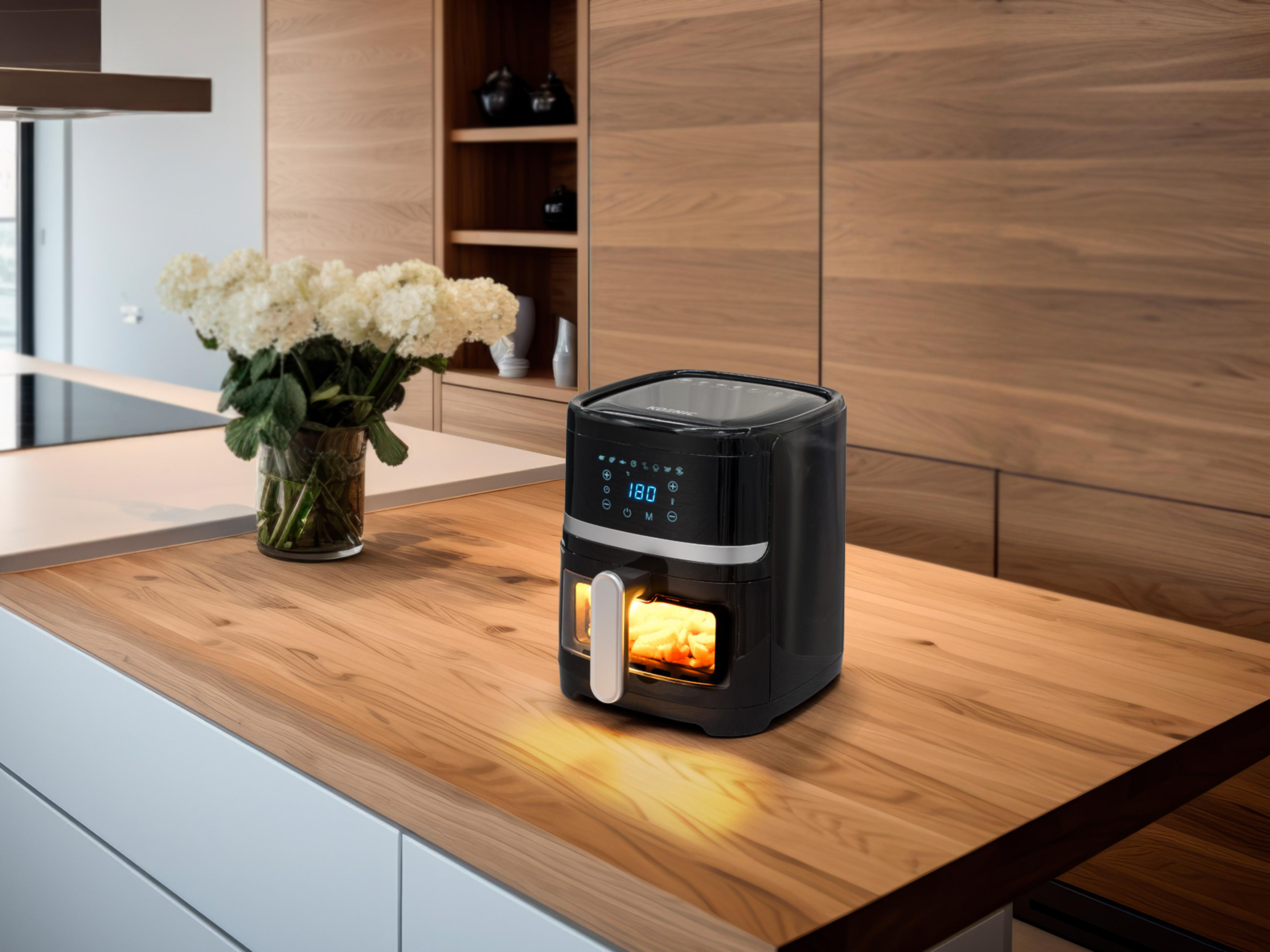 Schwarze Heißluftfritteuse auf einer Holztheke. Weiße Blumen und ein Holzschrank im Hintergrund.
