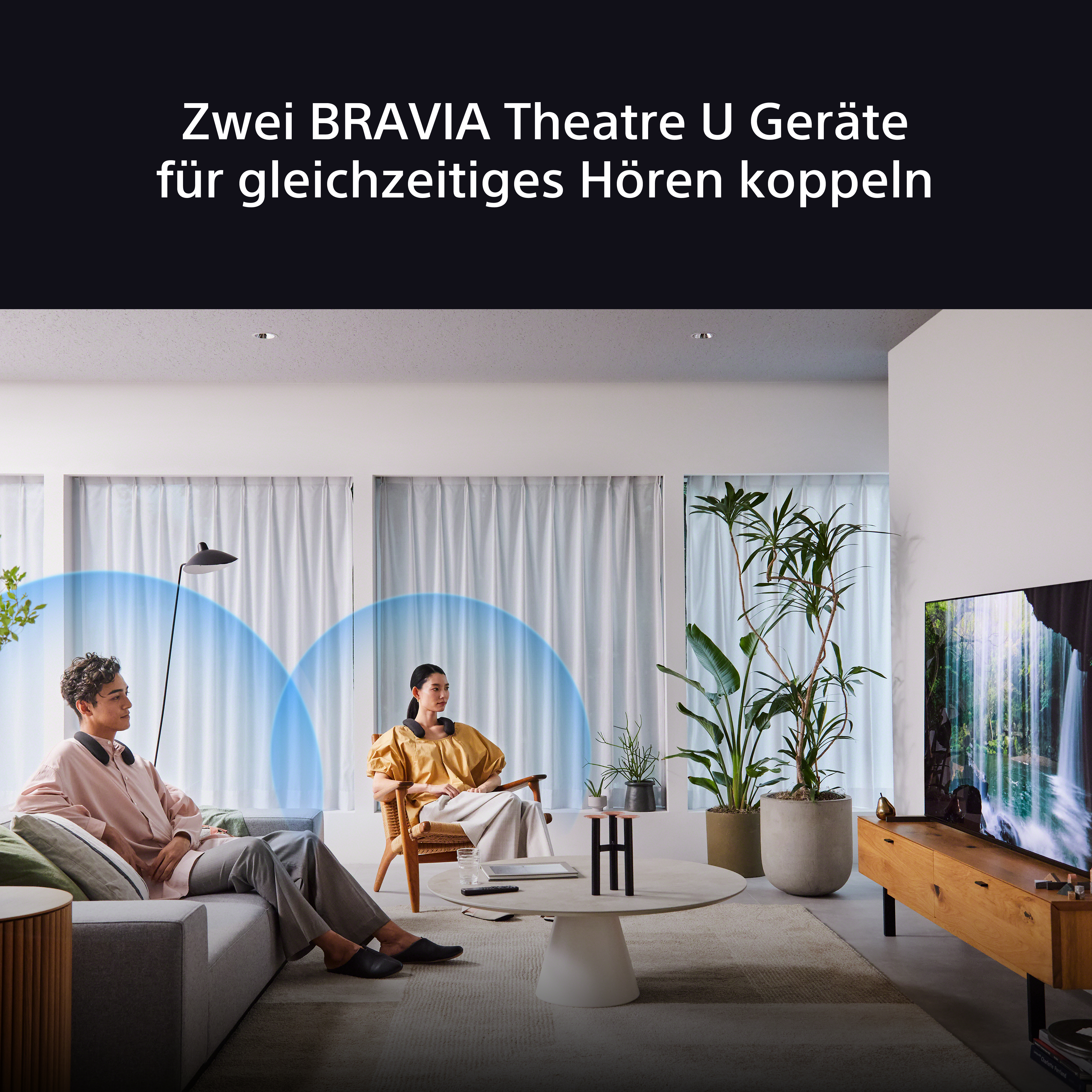 Zwei Personen mit Kopfhörern sitzen in einem Wohnzimmer und sehen fern.