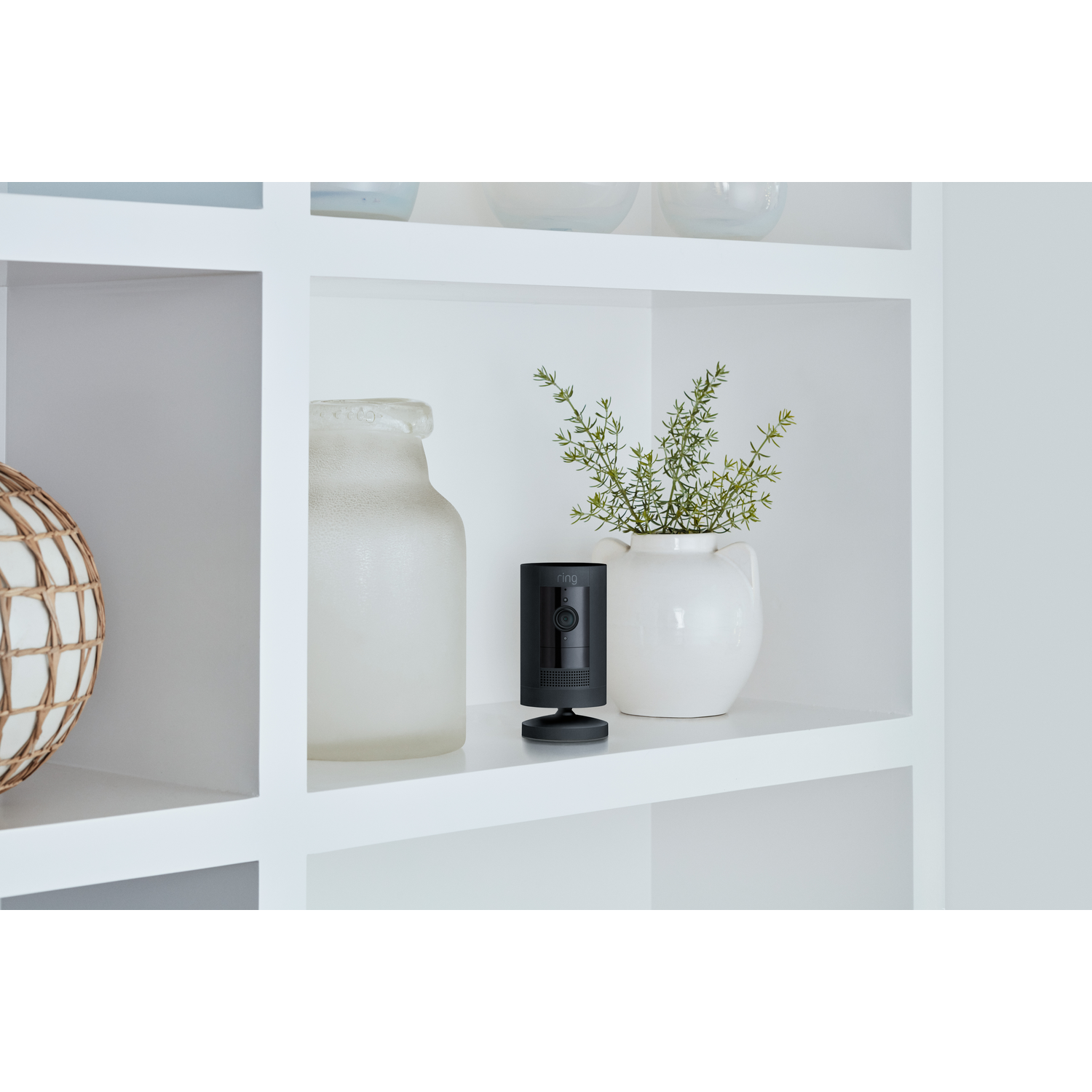 Black security camera on a shelf. Vase with plant, and decorative object nearby.