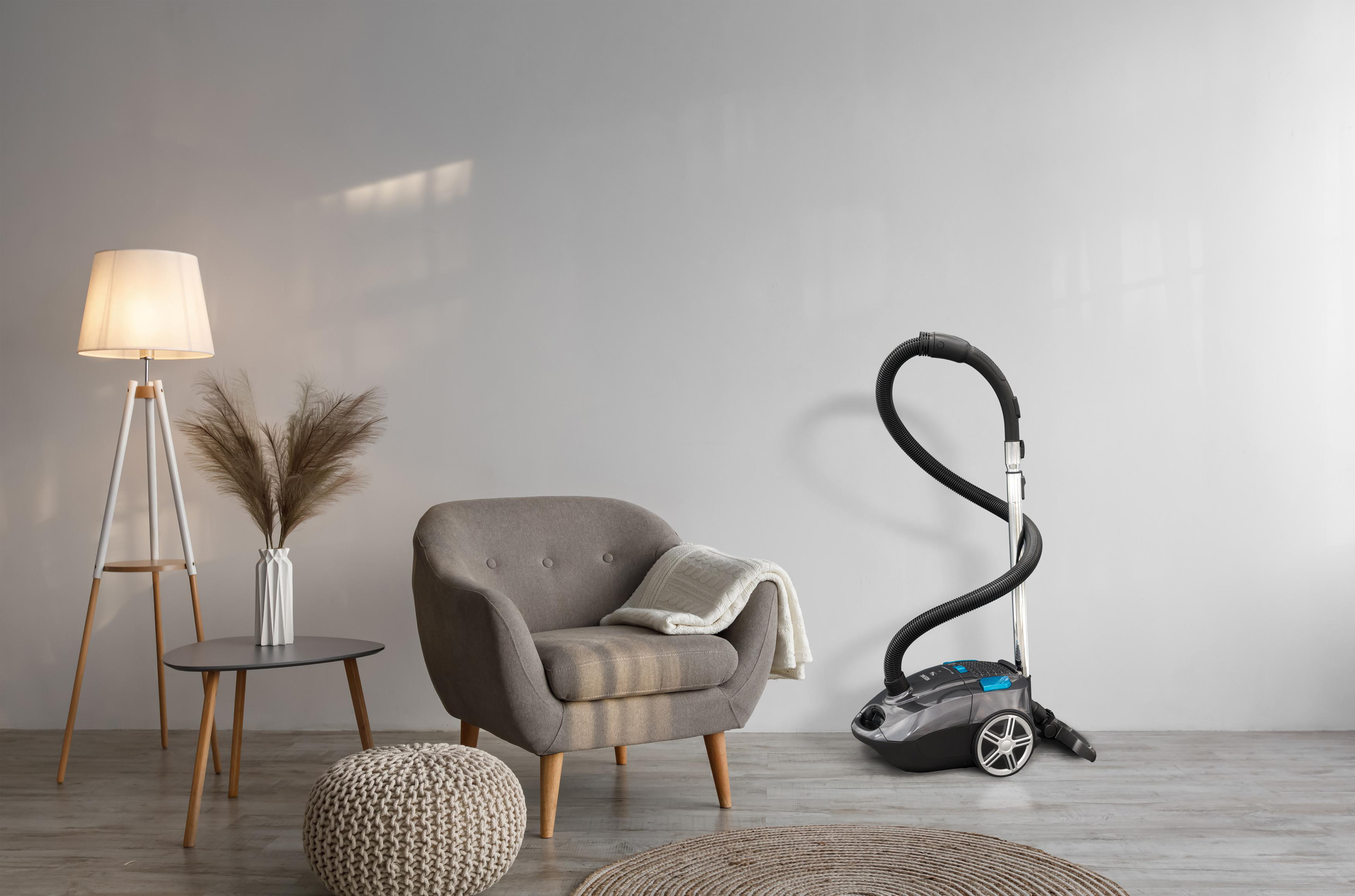 Zimmer mit grauem Sessel, Lampe und Staubsauger. Ein Teppich und ein Beistelltisch mit Pflanze.