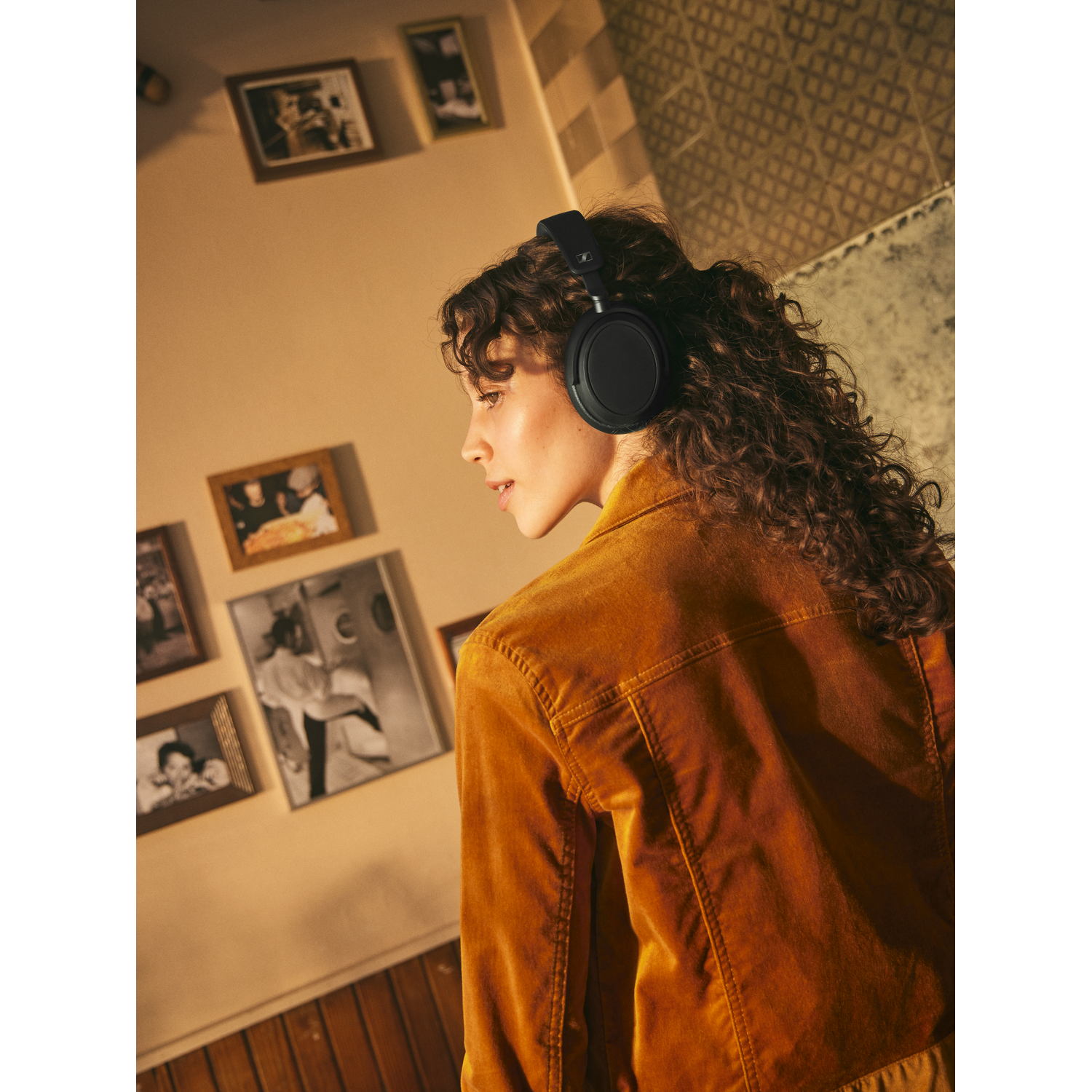 A woman wears black headphones and looks to the side. Photos on the wall in the background.