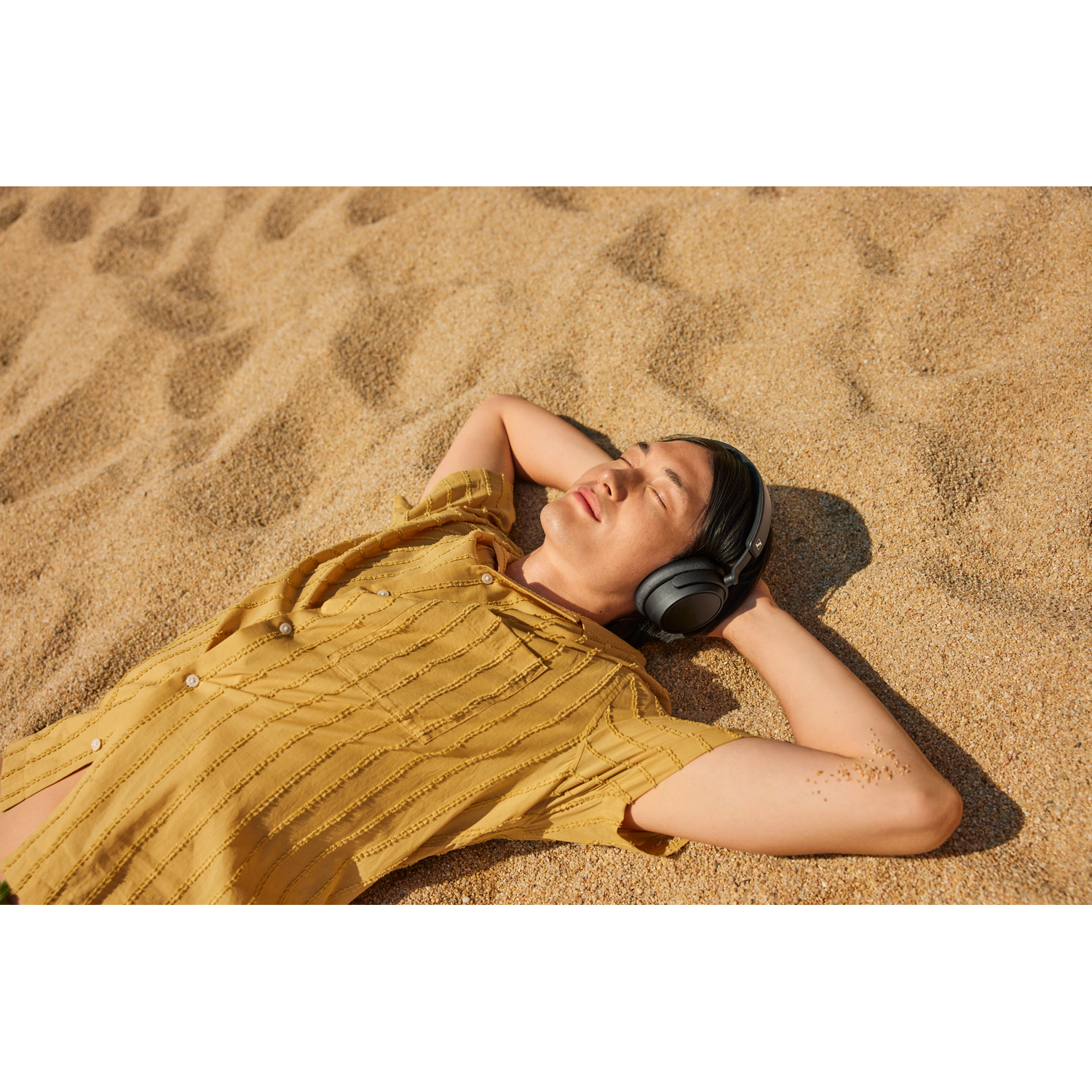 A man lies on a sandy beach wearing headphones, eyes closed, arms behind head.