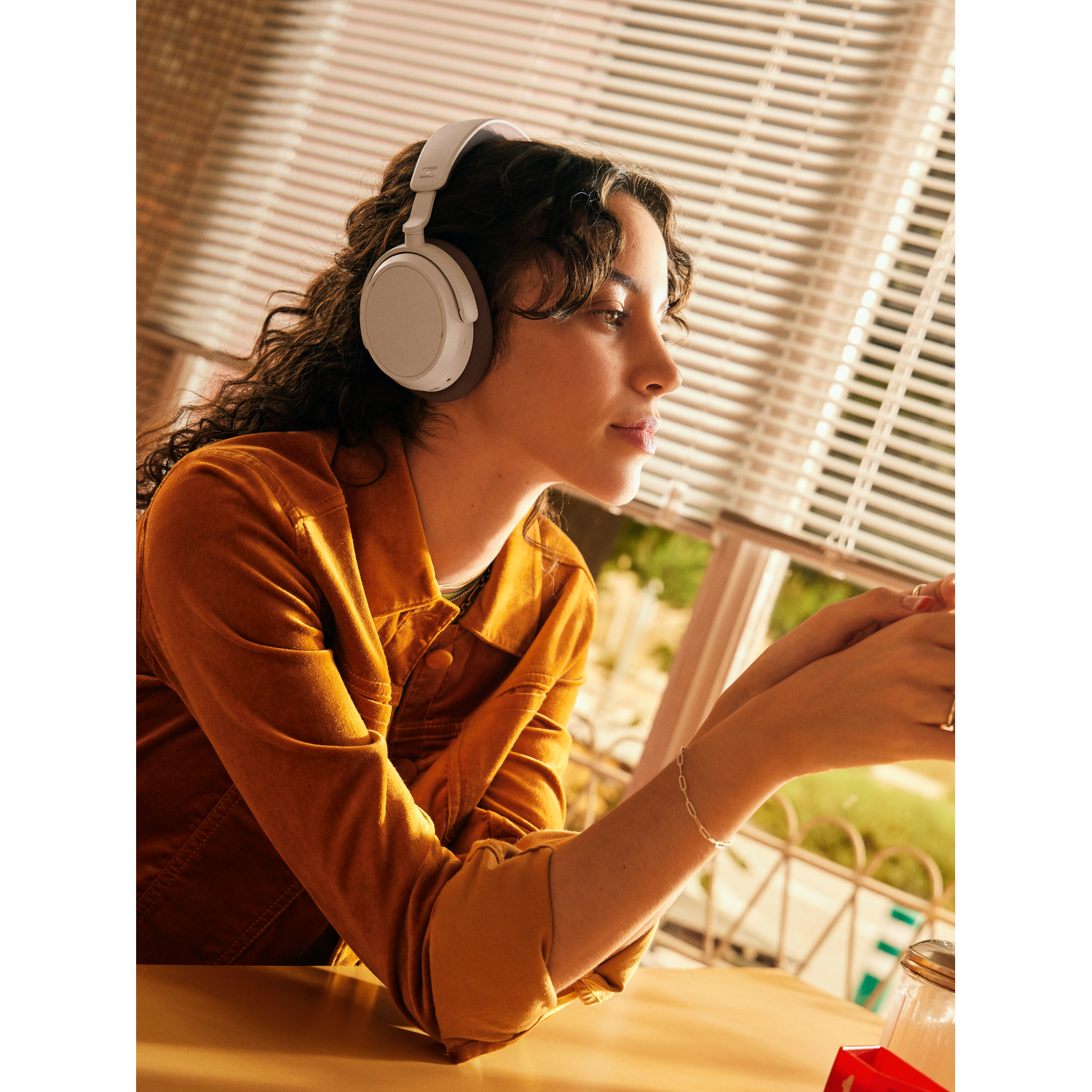 A woman with headphones sits near a window. She wears a tan jacket. The background shows a cafe setting.