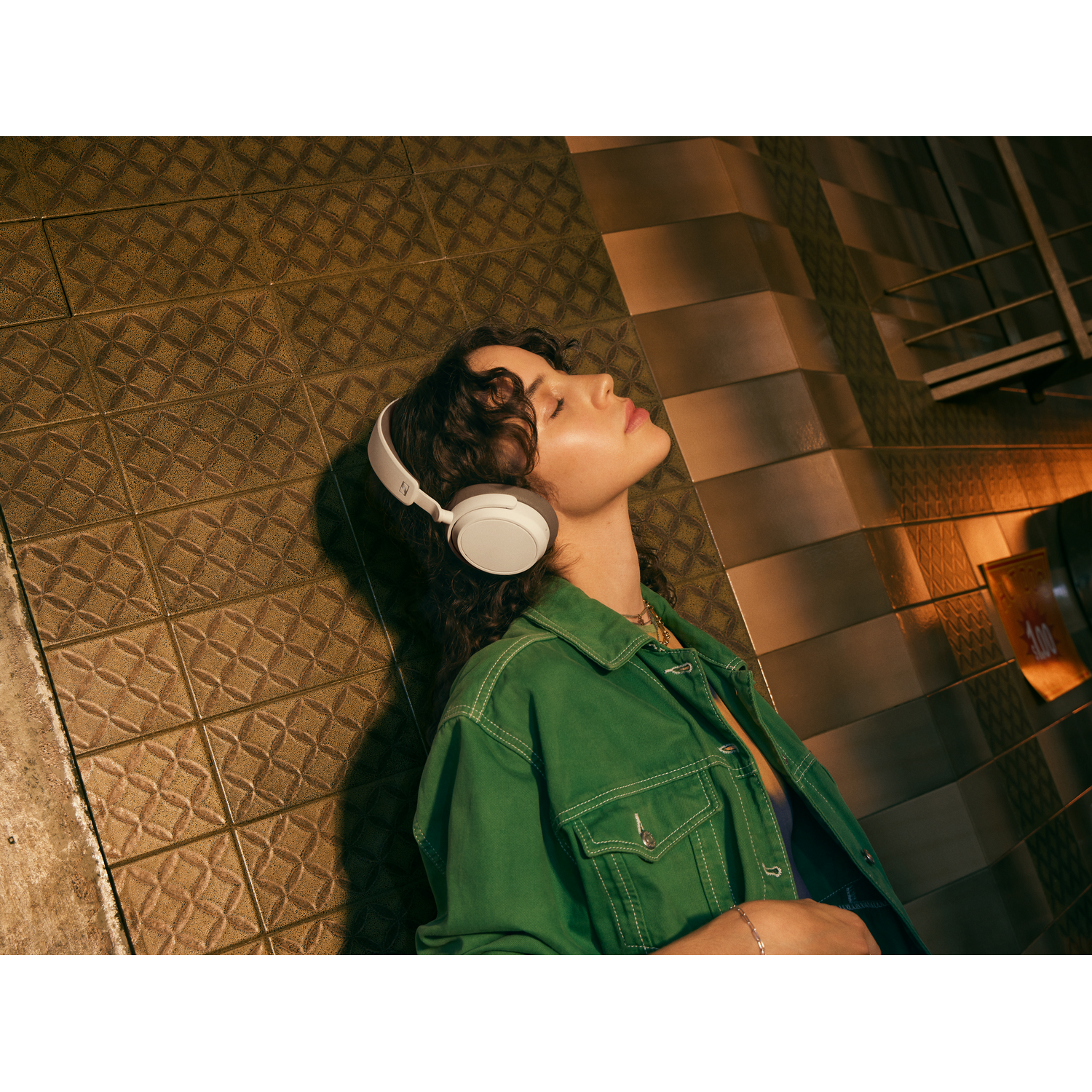 A woman with white headphones. She is leaning against a tiled wall, eyes closed.