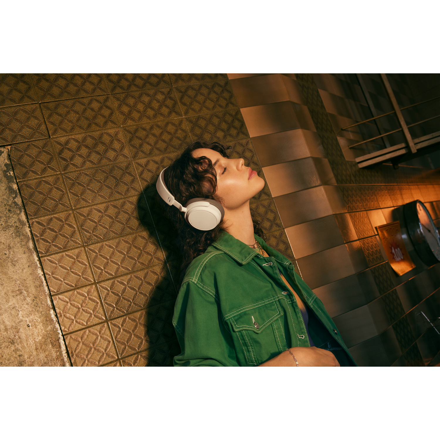 Woman wearing white headphones, looking up. Background is a tiled wall. Green jacket.