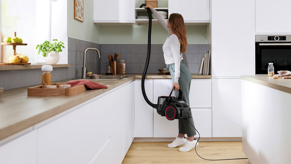 Beyaz bir mutfakta elektrik süpürgesi yapan bir kadın. Bir dolaba uzanıyor.