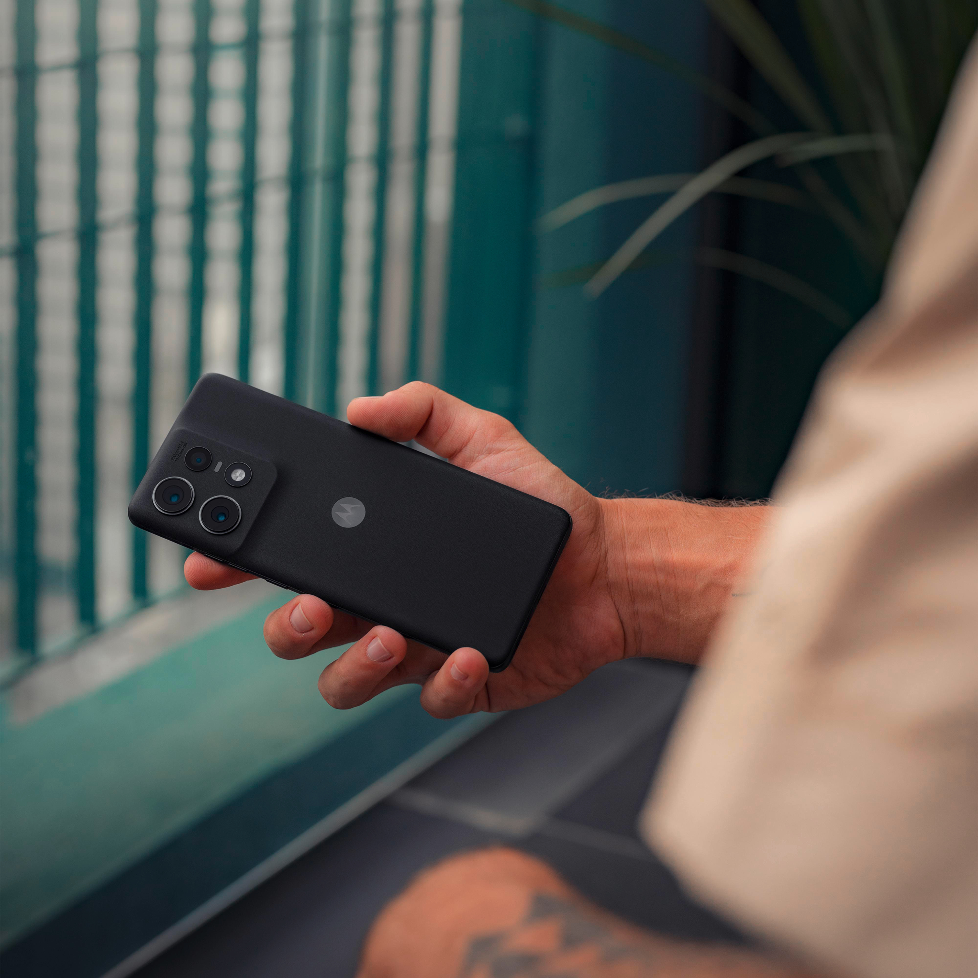 Person hält ein schwarzes Smartphone. Hintergrund ist eine grün-weiß gestreifte Wand.