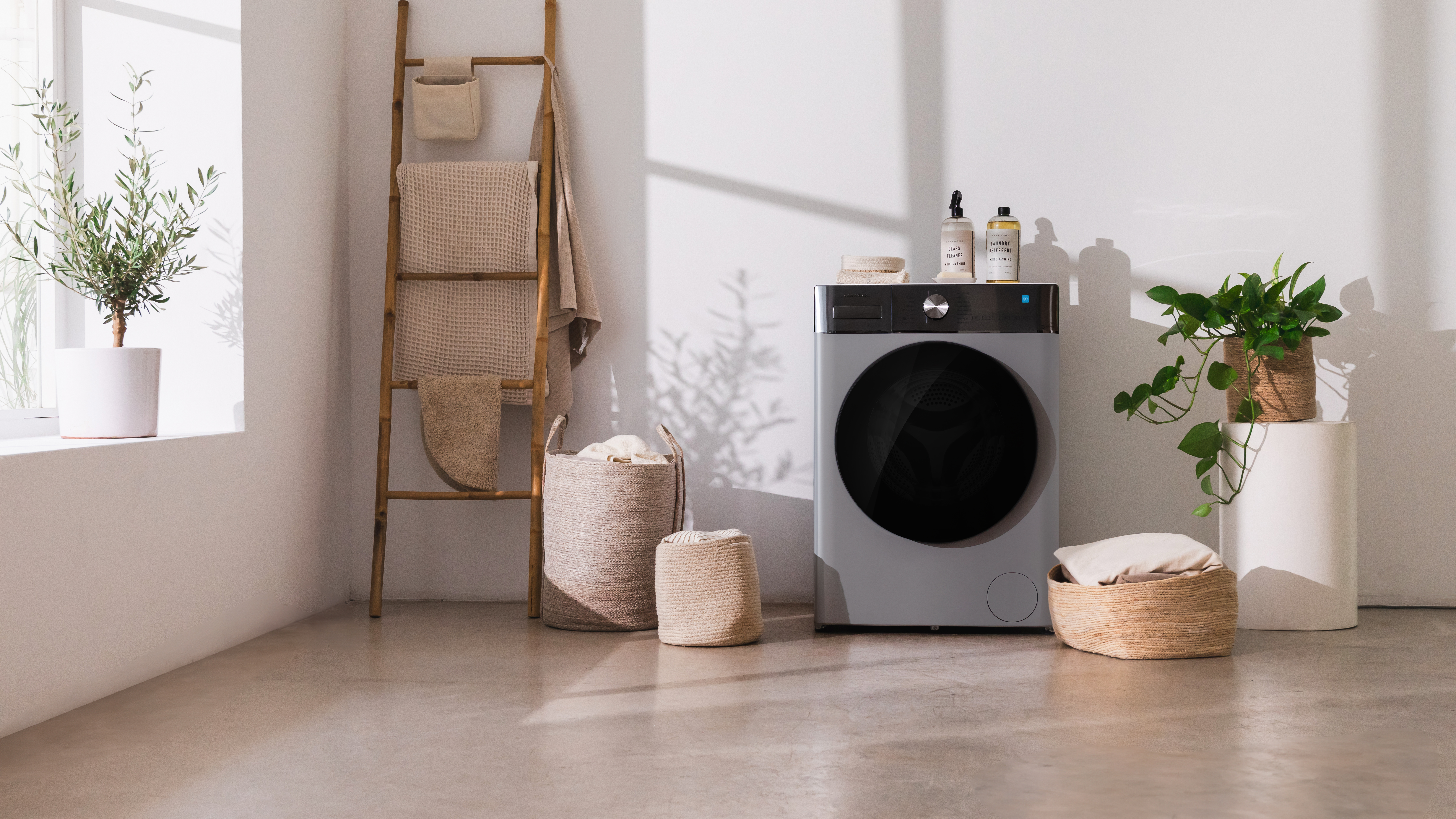Cuarto de lavado con lavadora, escalera de madera, cestas y plantas en macetas. Luz natural suave.