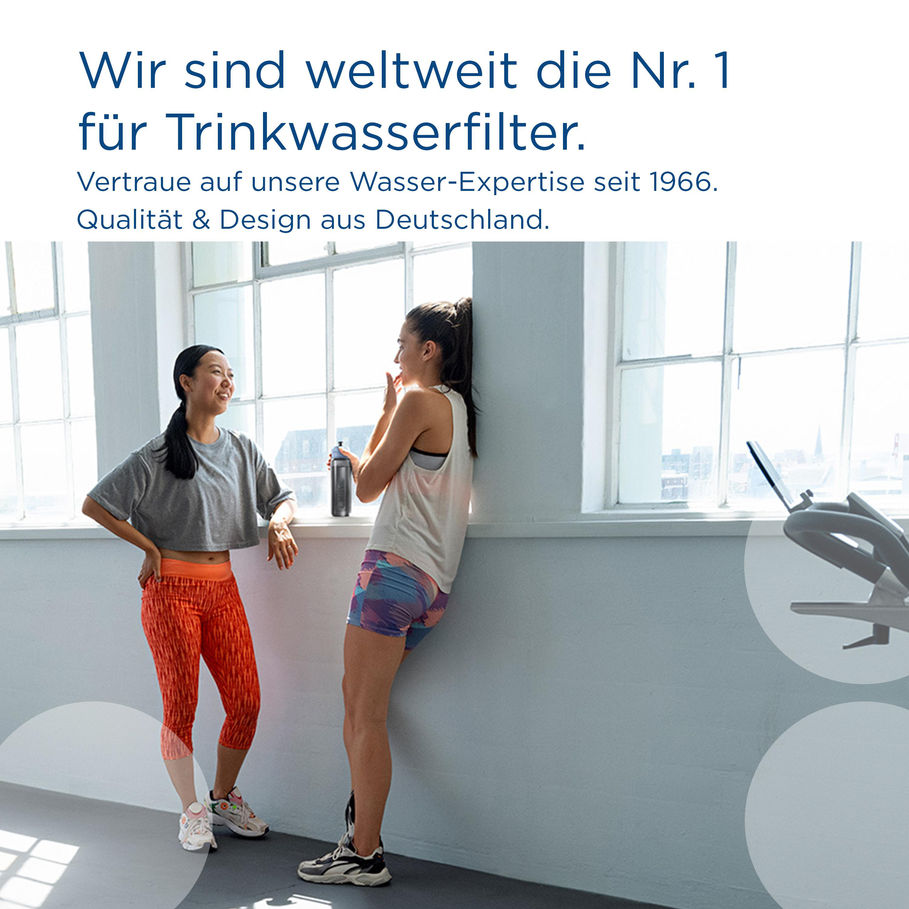 Zwei Frauen in einem hellen Raum, eine steht, lehnt an einer Wand, die andere spricht.