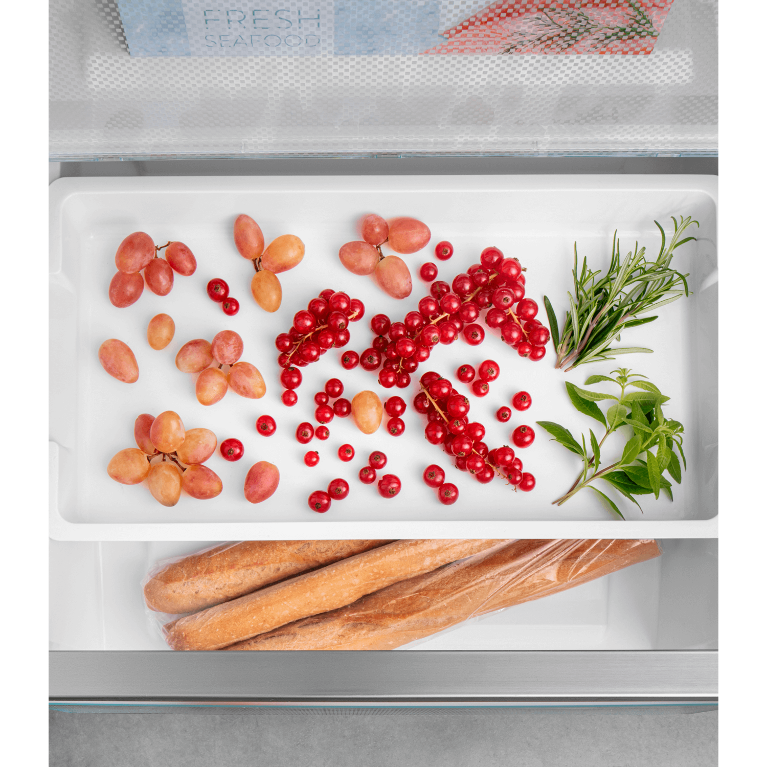 A refrigerator drawer filled with fresh produce: grapes, red currants, rosemary, and basil.