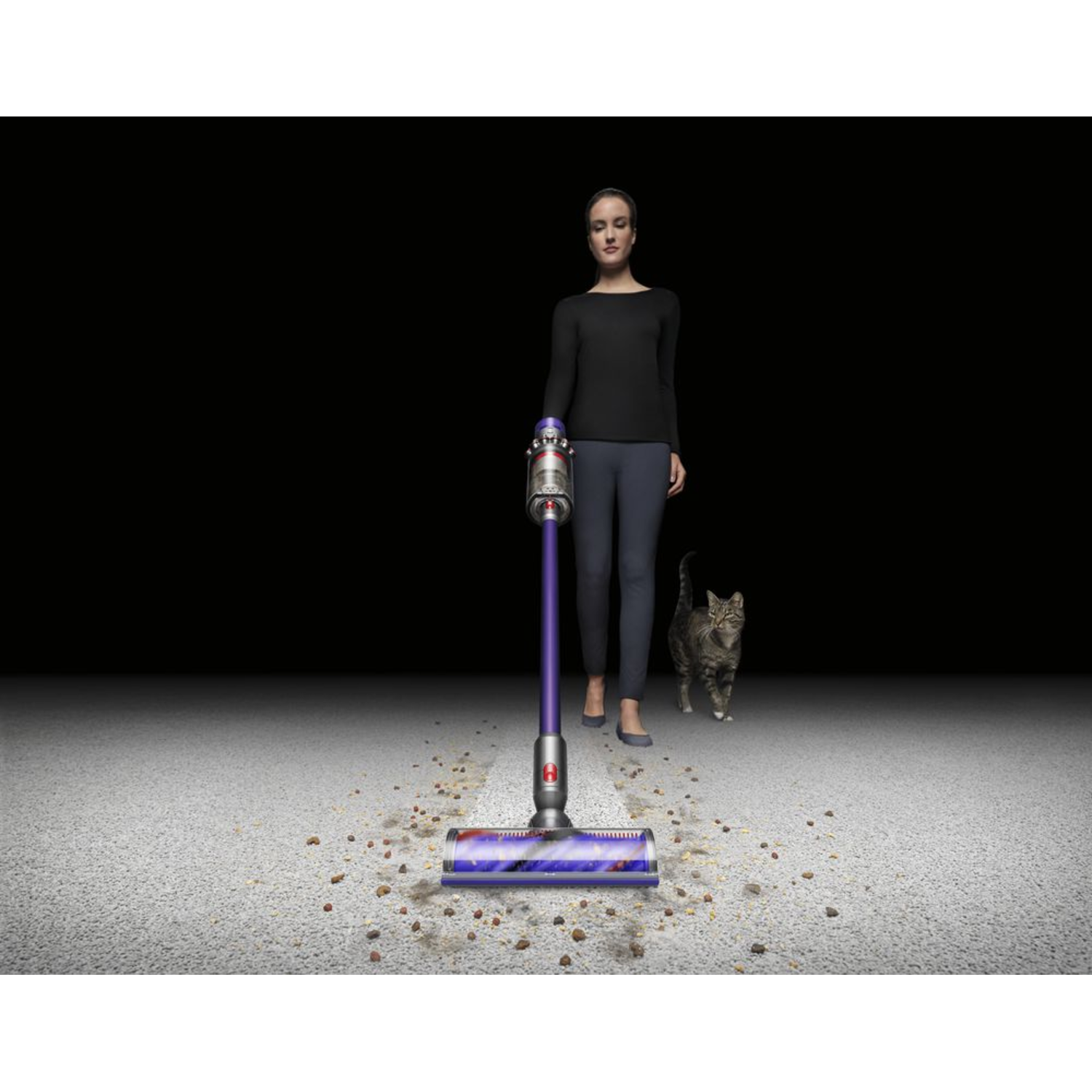Woman vacuums, cat watches, dirt on the floor. Black background, carpeted floor.